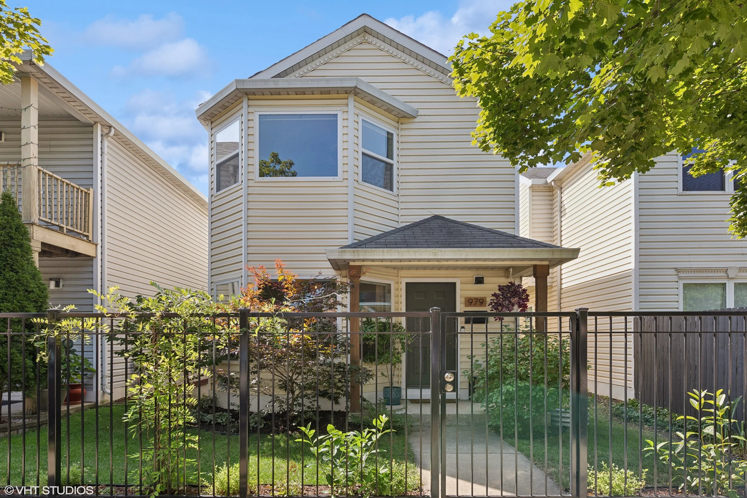 The front of a two-story house with cream-colored vinyl siding, a small porch, and a black metal fence in the foreground. A large tree with green leaves partially obscures the house, and the sky is blue with some clouds.