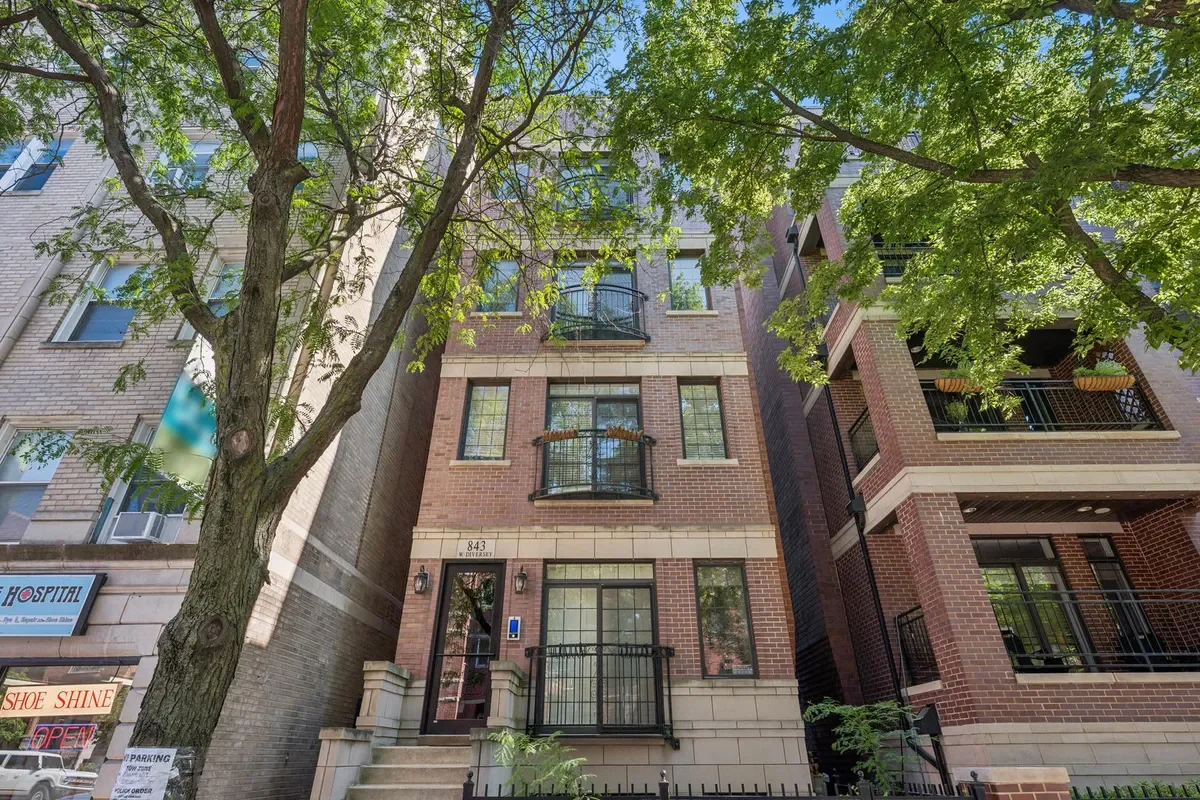 A multi-story brick residential building with black balconies, trees in front, and neighboring structures.