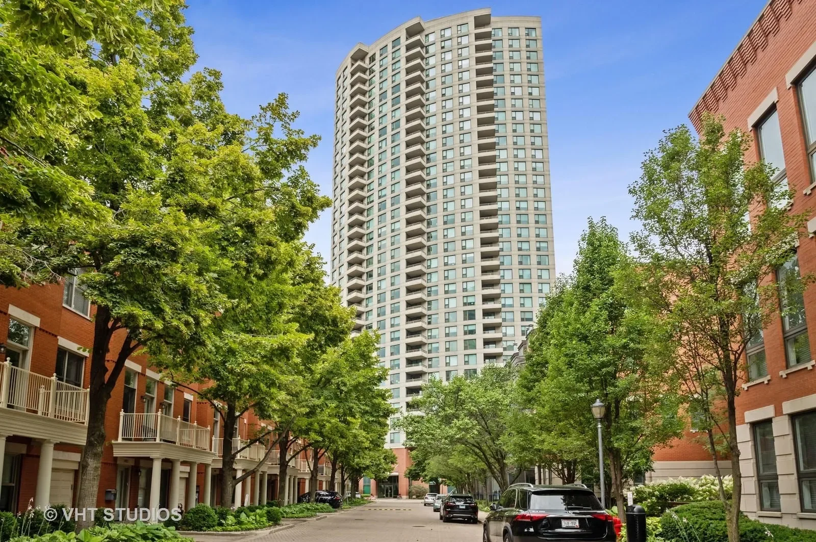A tall residential building with multiple balconies viewed from a street with lush green trees and parked cars, framed by a partly cloudy blue sky.