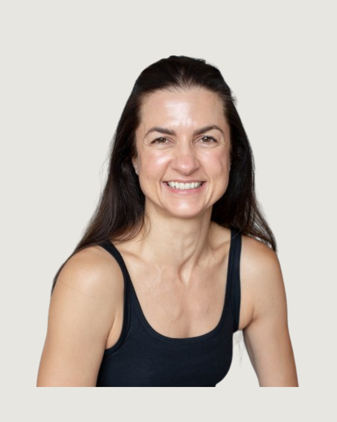 A woman with long dark hair, wearing a black tank top and light blue jeans, smiling at the camera in a bright room with white walls and hanging potted plants in the background.