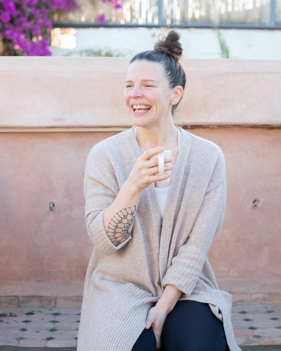 A woman with dark hair in a bun, wearing a beige cardigan, holding a white mug, smiling outdoors against a pinkish wall with purple flowers visible in the background.