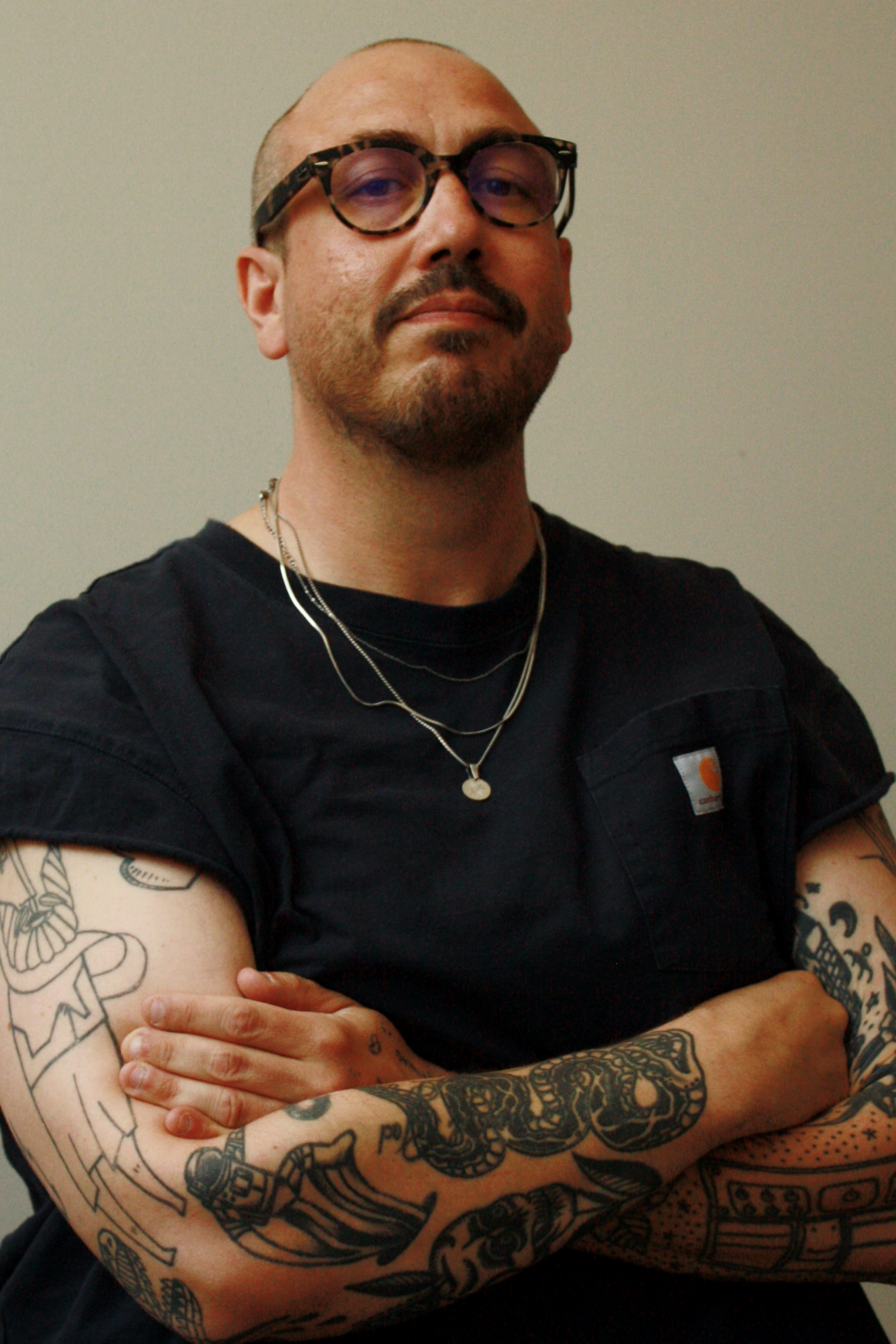Man with tattoos wearing glasses, black shirt, layered necklaces, with arms crossed, standing against a plain background.