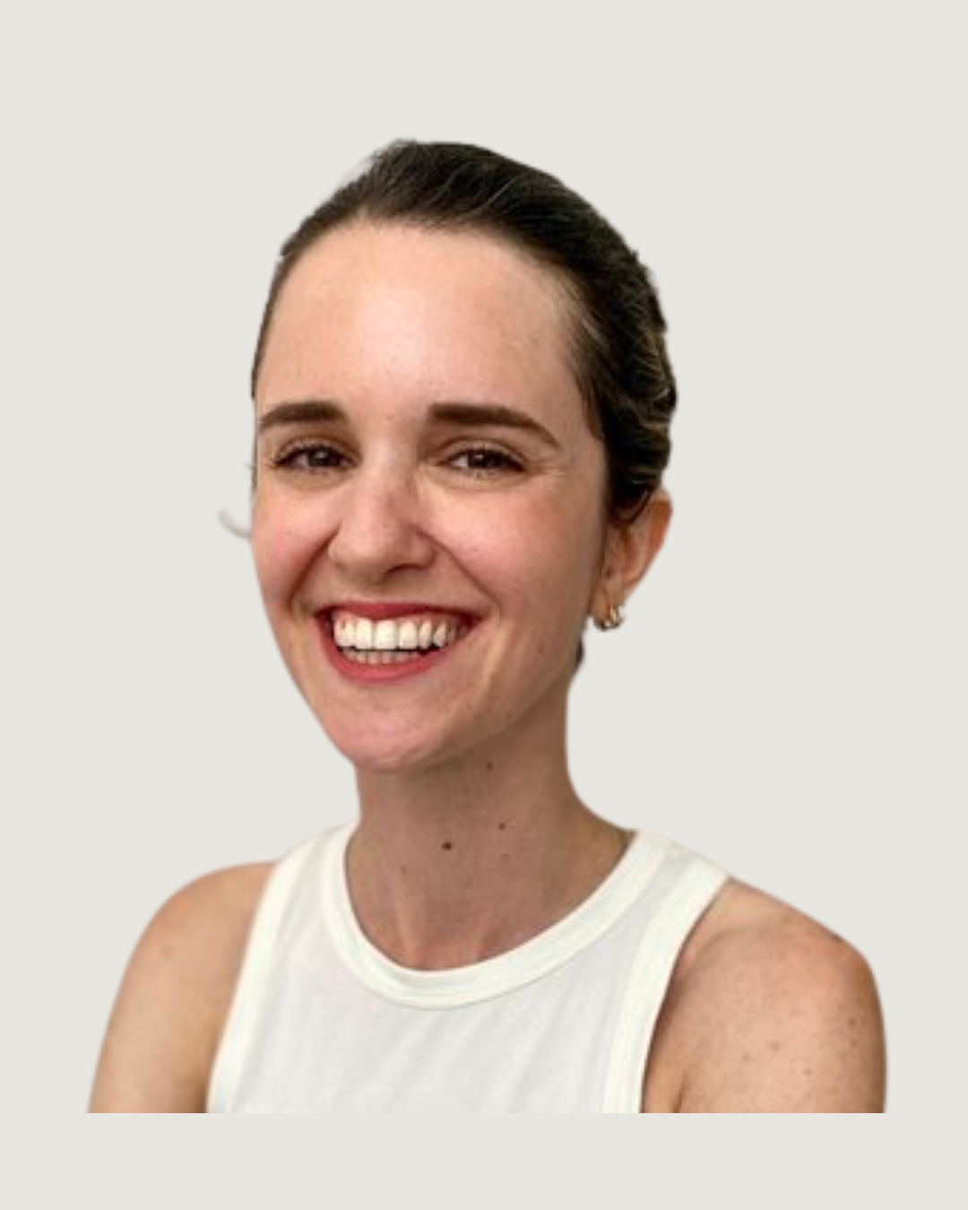 A smiling woman with brown hair tied back, wearing a sleeveless white top and earrings, standing with arms crossed against a plain light-colored wall.