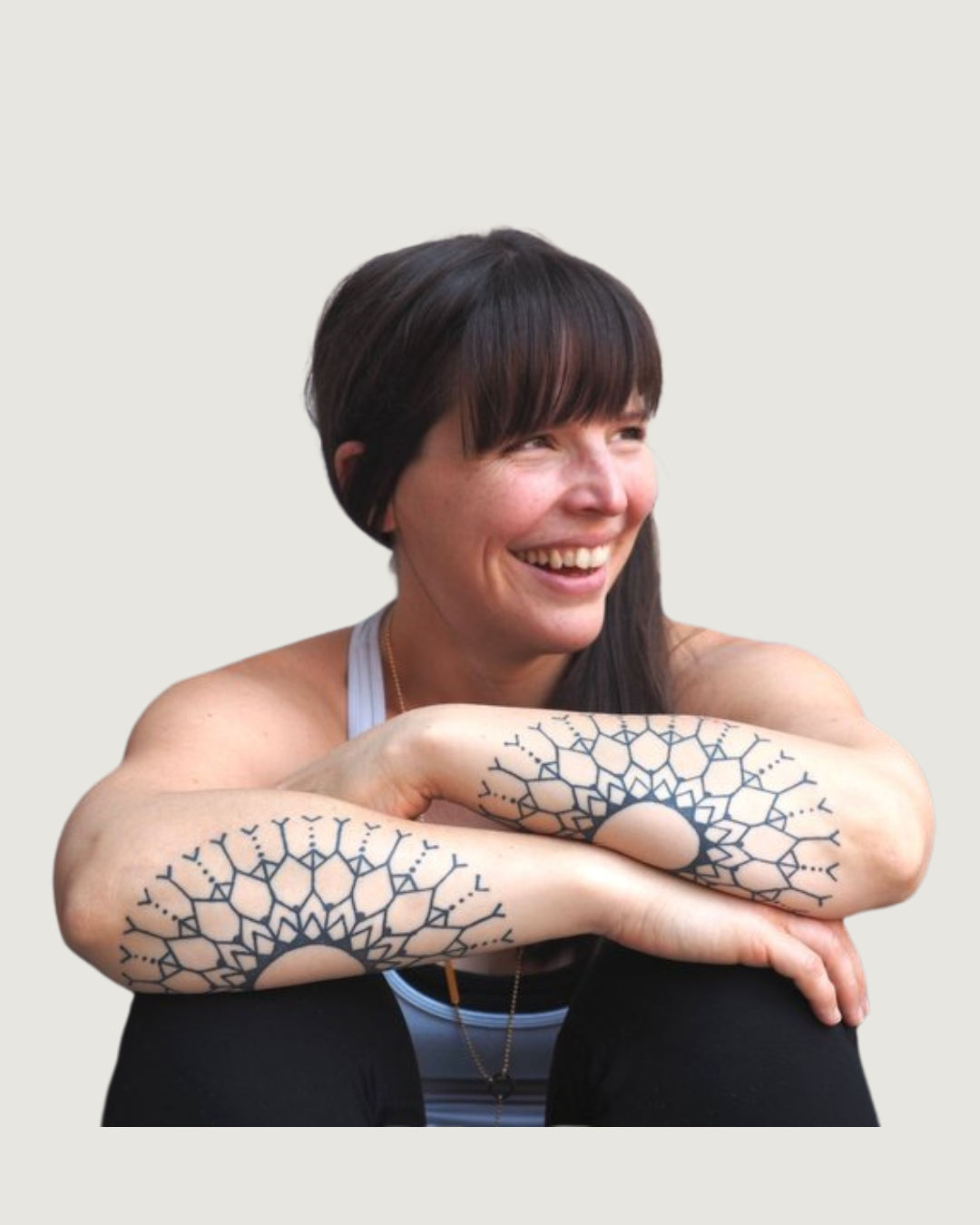 A smiling woman with dark brown hair and tattoos on her arms, sitting against a wall.