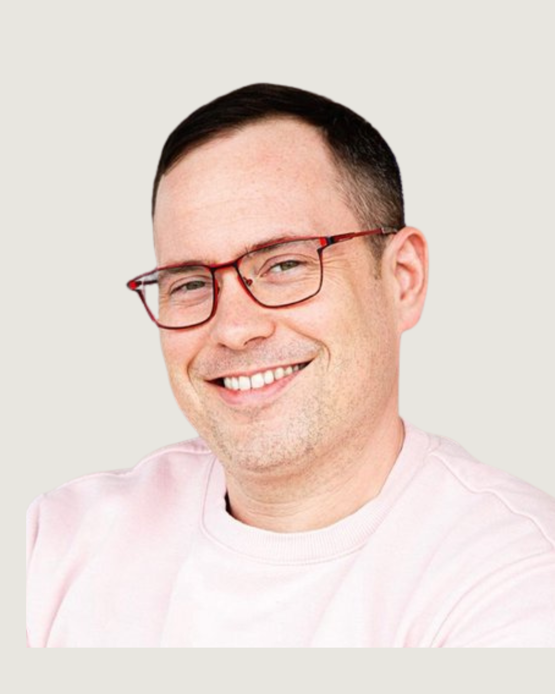 A smiling man with short dark hair, light skin, wearing red glasses and a pale pink shirt, indoors in a bright, modern setting.