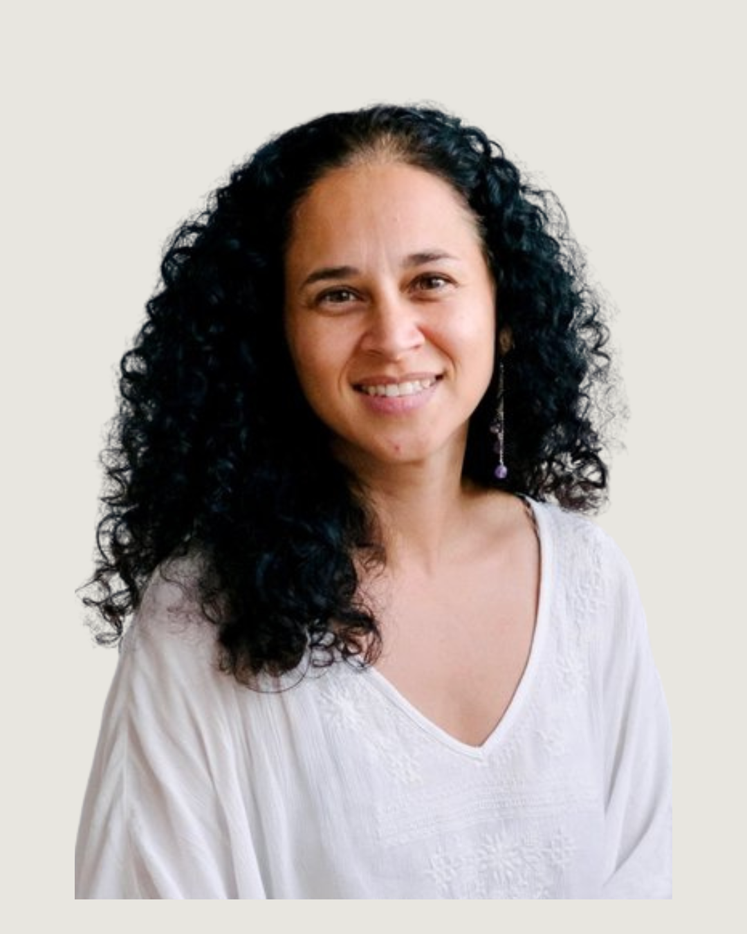A woman with curly dark hair smiling, wearing a white embroidered dress and earrings, sitting in front of a white paneled wall.
