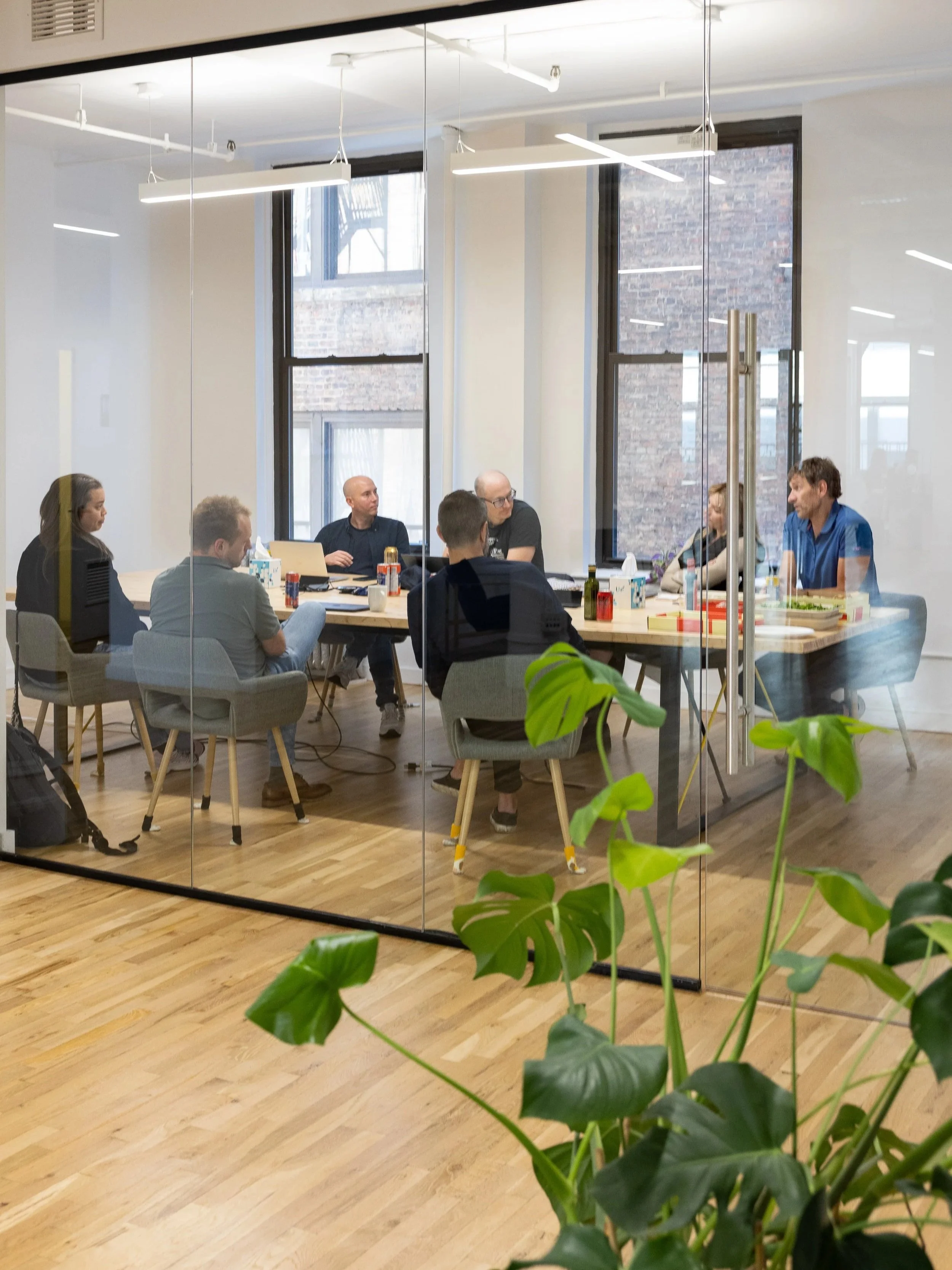 A group of seven people sitting around a conference table in a modern office, seen through a glass wall, with large windows and wood flooring, and a green plant in the foreground.