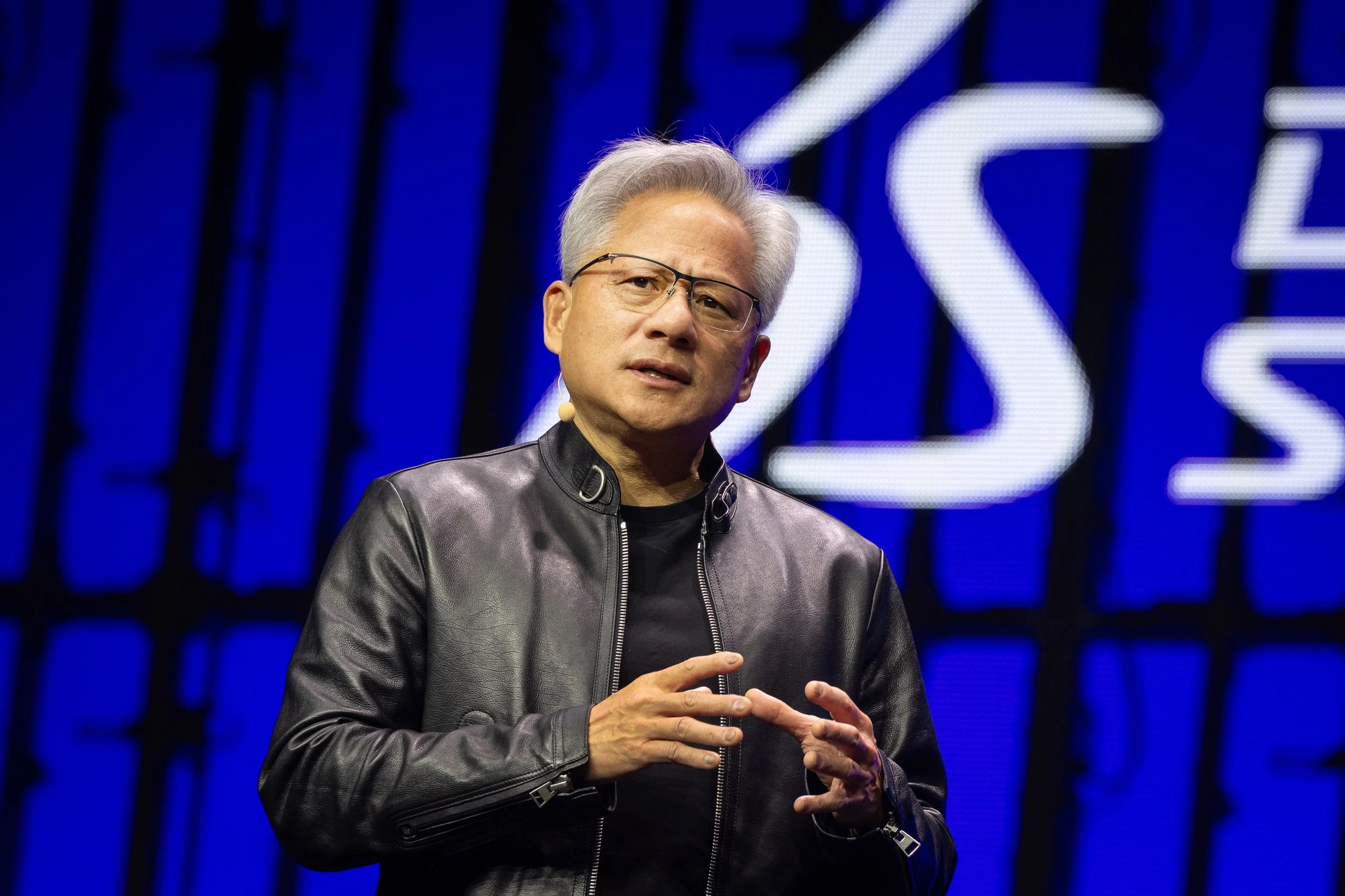 A man with gray hair and glasses speaking on stage, wearing a black leather jacket, with a blue background featuring large white illuminated letters.