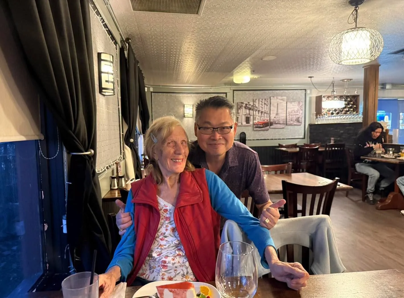 A smiling elderly woman and a man are seated at a restaurant table, posing for a photo. The woman is wearing a floral top with a red vest and blue cardigan, and the man is wearing glasses and a dark purple shirt. The table has a glass of water, a pla