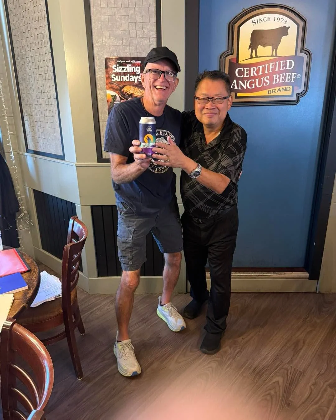 Two men smiling, one holding a can of colorful beverage, inside a restaurant with a sign that reads 'Certified Angus Beef' in the background.