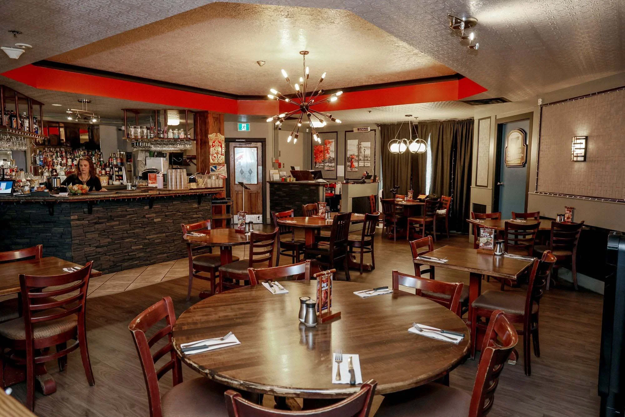 Interior of a restaurant with wooden tables set with napkins, forks, knives, and salt and pepper shakers, and empty chairs surrounding the tables. There is a bar area with bottles and glasses behind the woman serving behind the bar, and decorative lighting fixtures hang from the ceiling.