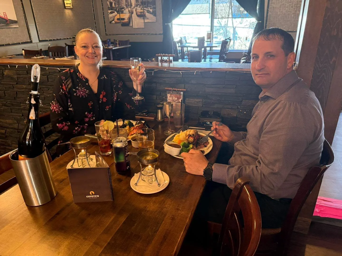 A man and woman dining at a restaurant table with food and drinks, the woman holding a glass of wine, the man eating a meal, on a wooden table with a bottle of wine, glasses, and small plates, in a cozy restaurant setting.