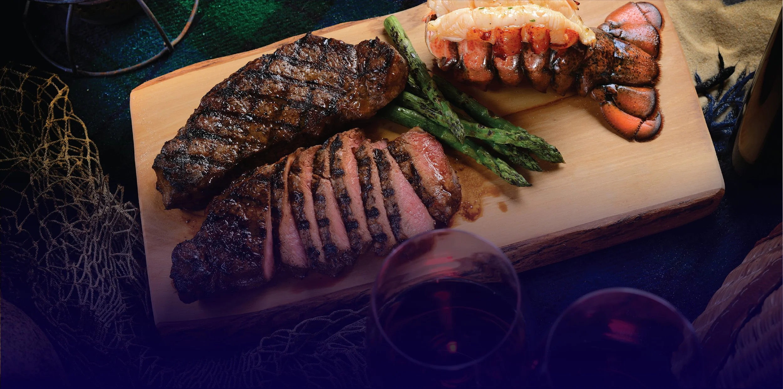 A wooden serving board with a grilled steak, sliced and cooked to medium rare, grilled asparagus, and a lobster tail. Two glasses of red wine are visible in the foreground.