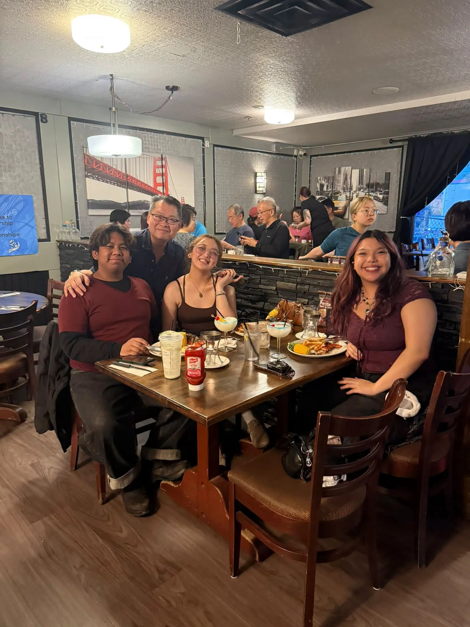 People dining at a restaurant table with food and drinks, including cocktails, with a cityscape artwork and gray walls in the background.