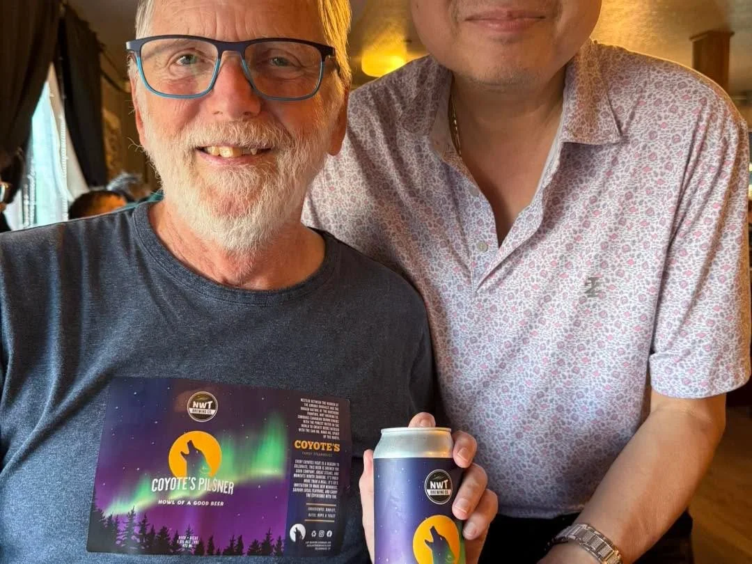 Two men, one with glasses and a beard holding a can of beer and a promotional card, smiling at the camera inside a restaurant or bar with warm lighting.
