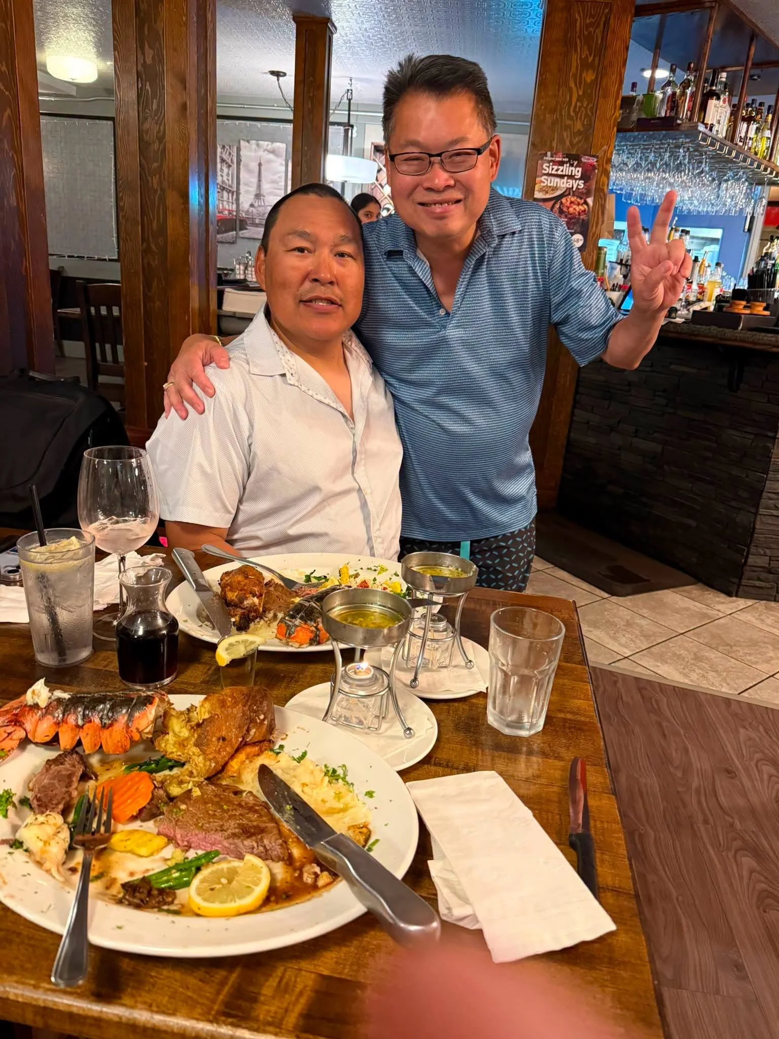 Two men celebrating at a restaurant, with a large plate of assorted foods including seafood, steak, and vegetables on the table, along with glasses of water and wine.