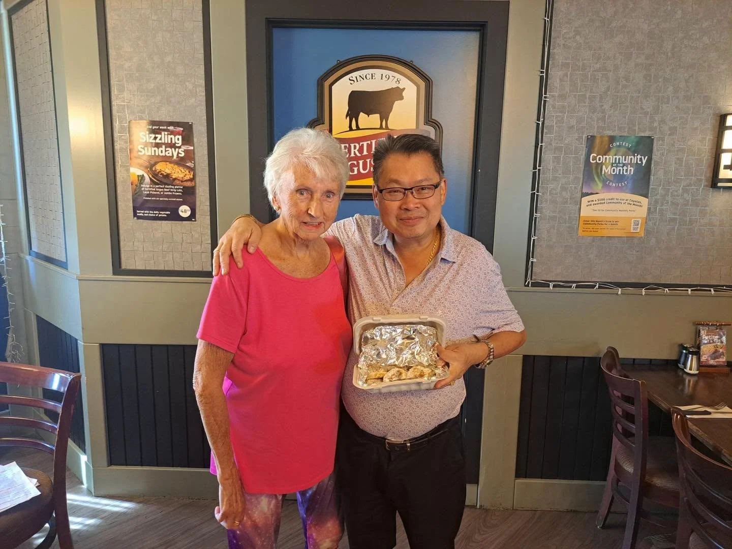 An elderly woman and a man are standing together inside a restaurant, smiling at the camera. The man is holding a takeout container with food. Behind them, a sign on the wall features a cow silhouette and the text 'Since 1978'. The restaurant interio