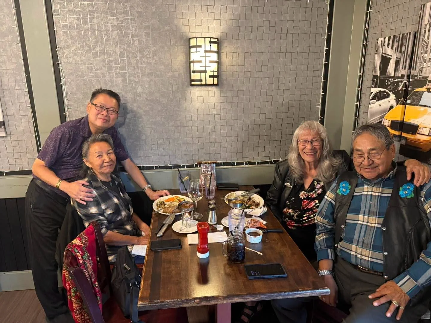 A group of five people, two women and three men, gathered around a dinner table in a restaurant. They are smiling and appear to be enjoying a meal together. The table has plates of food, drinks, and utensils on it.
