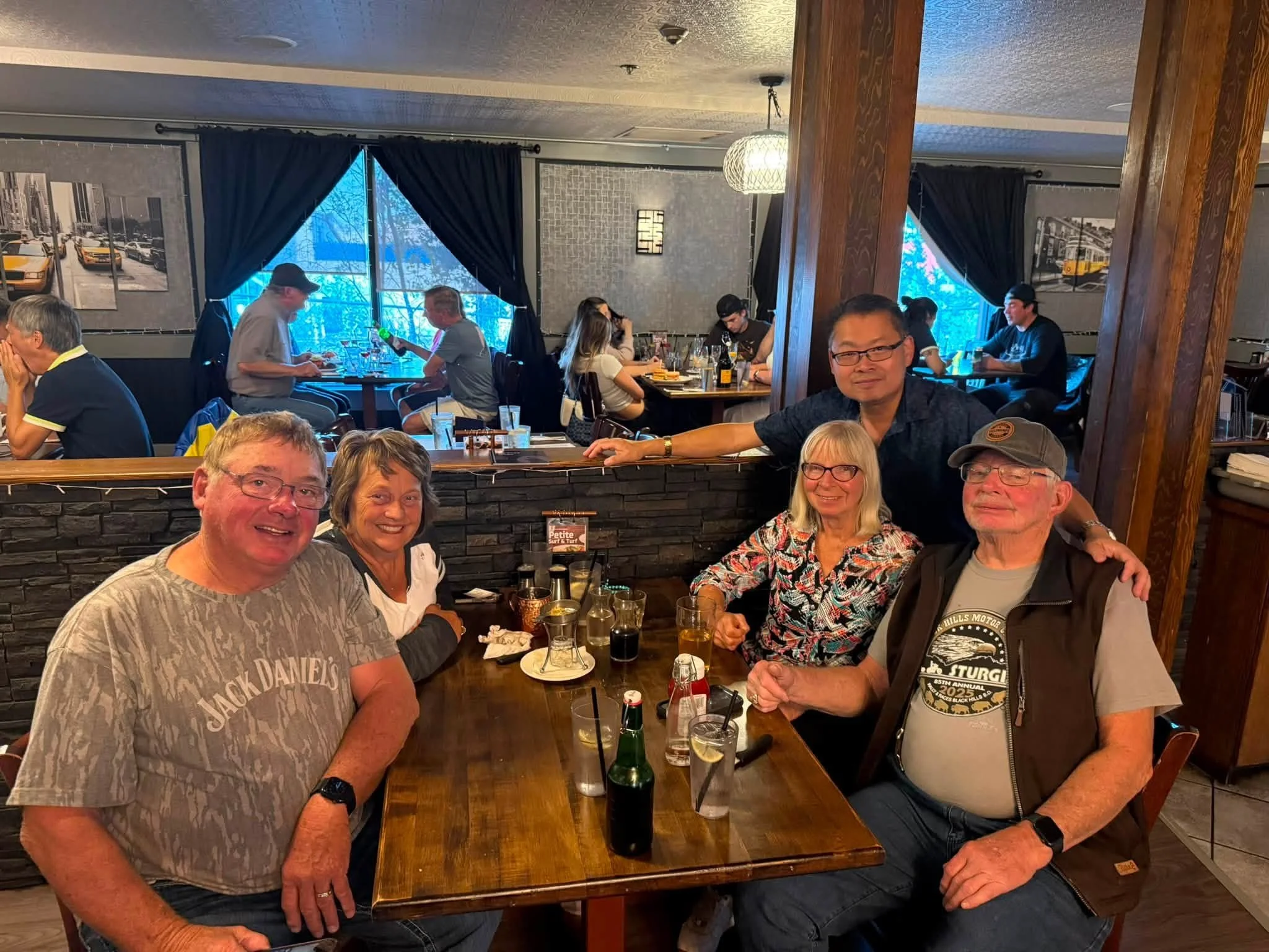 A group of older adults sitting around a wooden table in a restaurant, smiling and enjoying drinks. The background shows more patrons, large windows with black curtains, and framed black-and-white cityscape photos on the walls.
