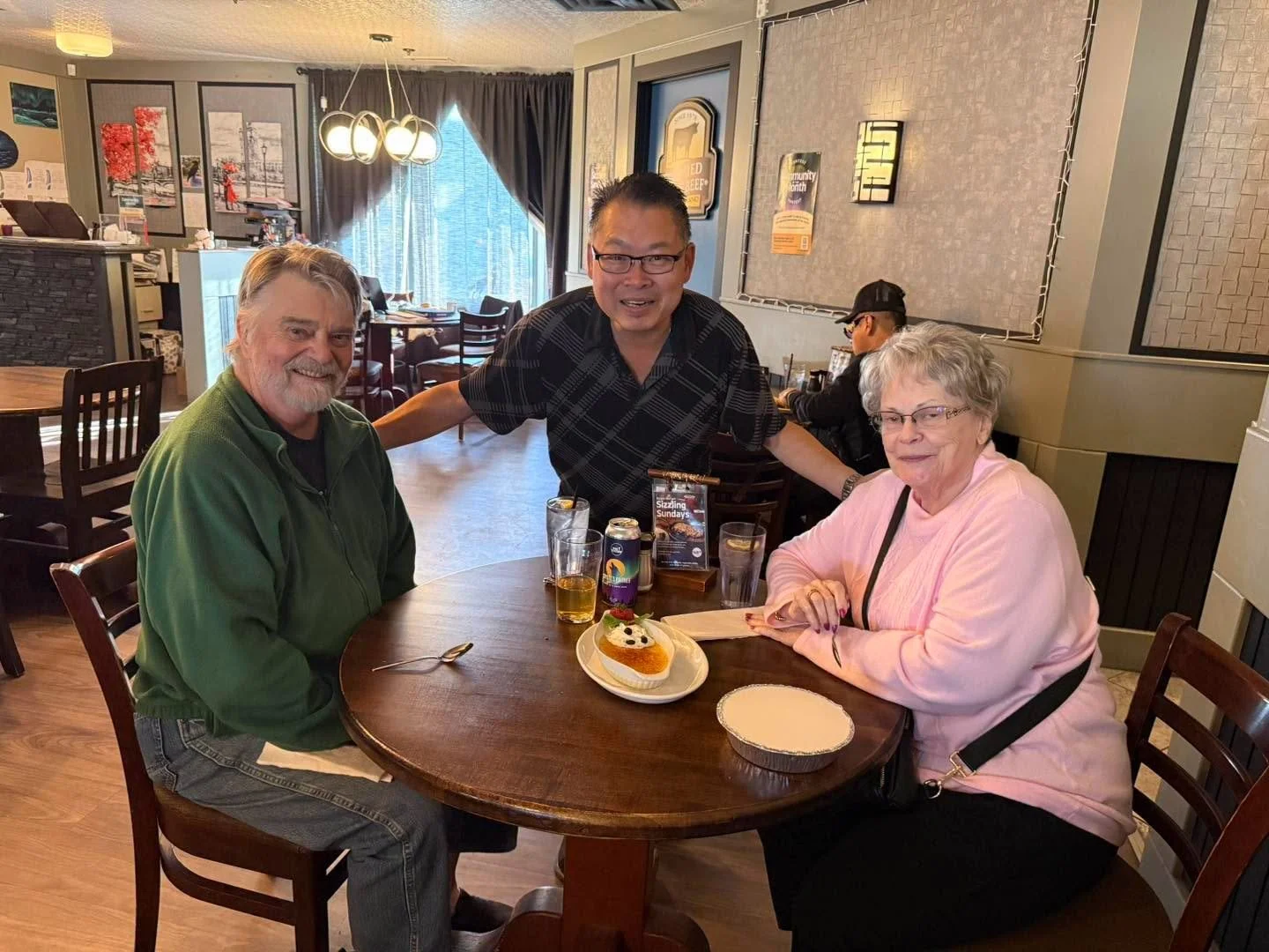 Three people sitting around a table in a restaurant with a cake and drinks, smiling for the photo.