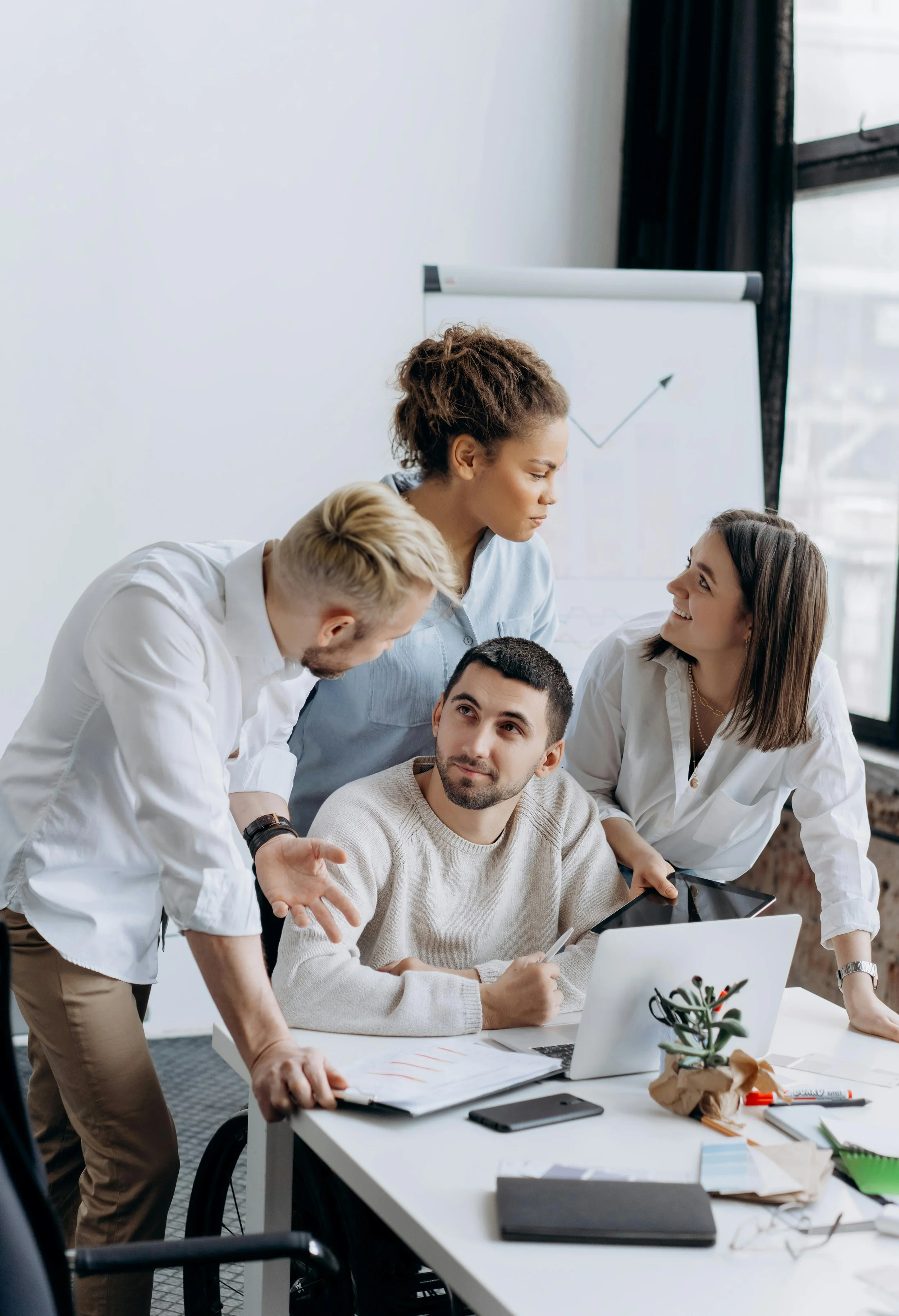 Four diverse colleagues in a business meeting discussing around a table with open laptops and documents, with a whiteboard showing a rising graph in the background.