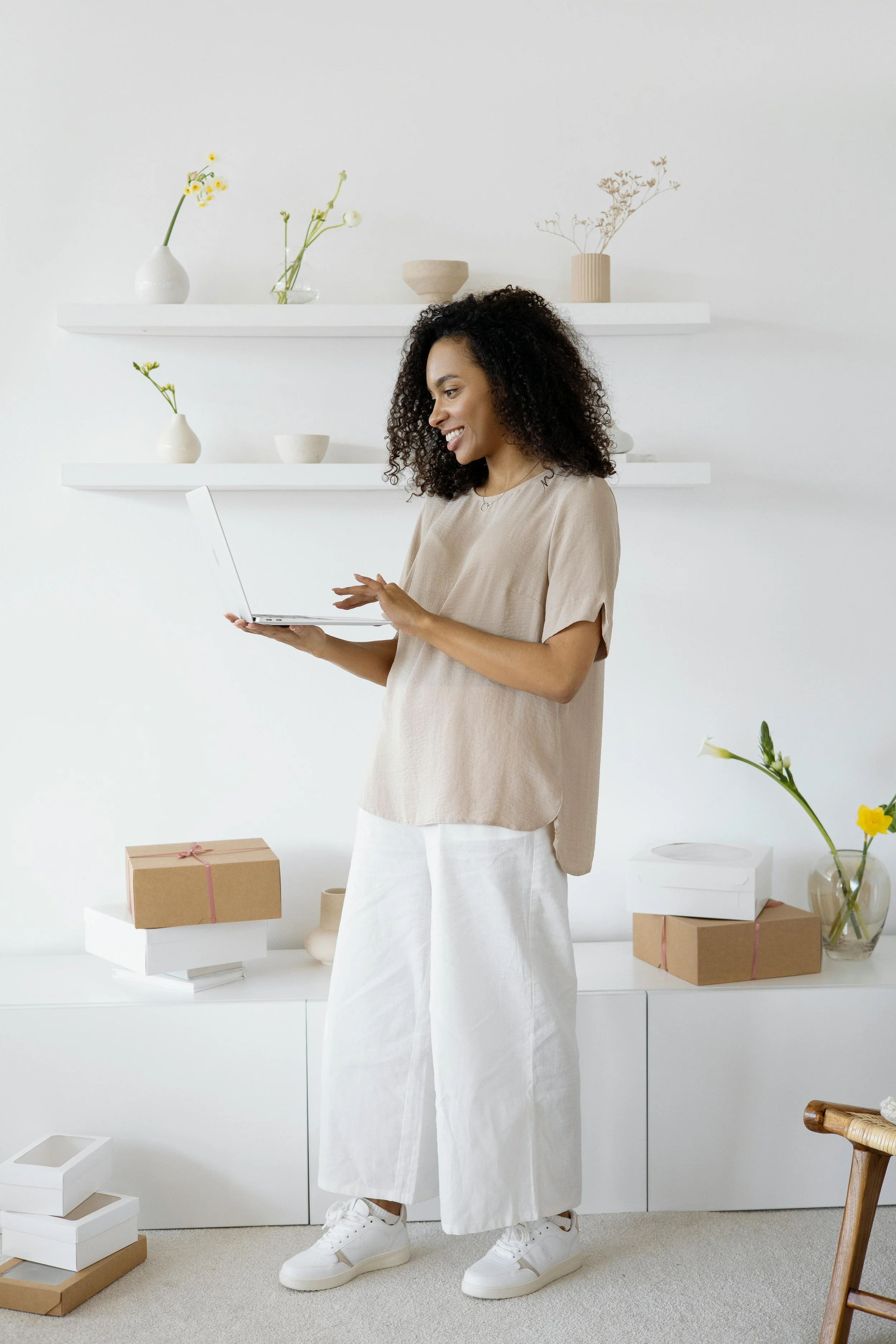 A woman with curly hair smiling while looking at her laptop in a minimalist, white-decorated room with shelves, vases, and gift boxes.