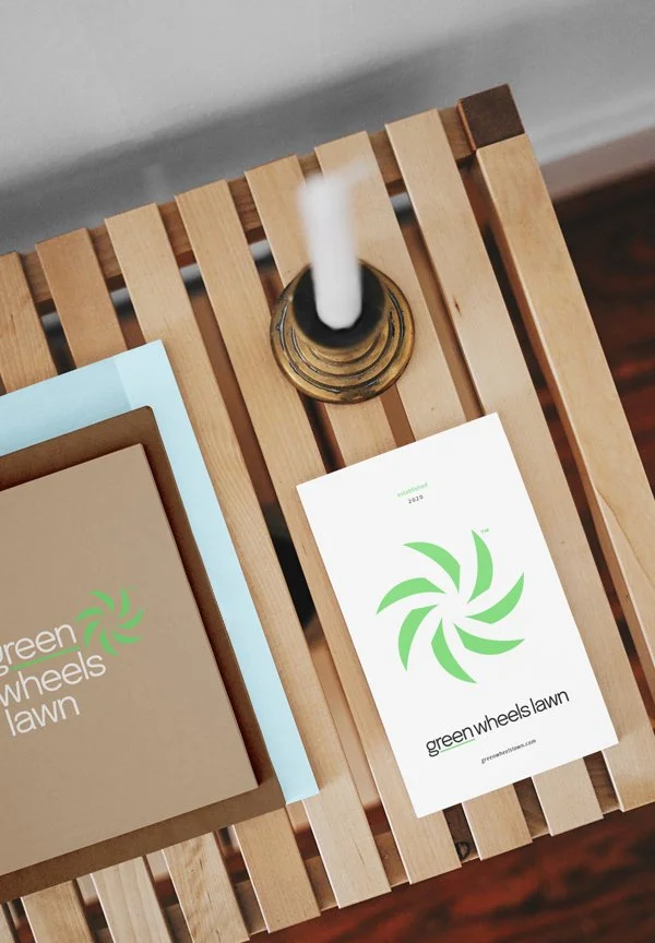 A wooden side table with a white candle in a brass holder, and promotional materials for 'green wheels lawn,' including a brochure and a card with a green swirling logo.