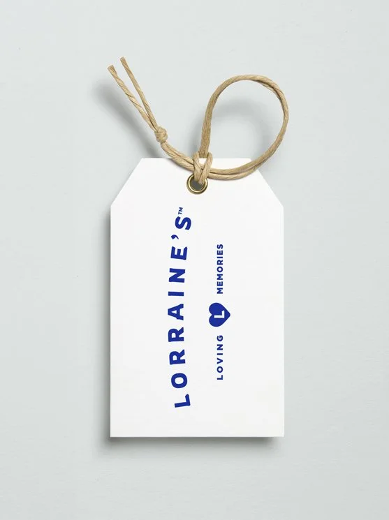 Designed by Infinite® - White gift tag with blue text that reads "LORRAINE'S LOVING MEMORIES" branding and a small heart symbol, attached to a string on a white background.