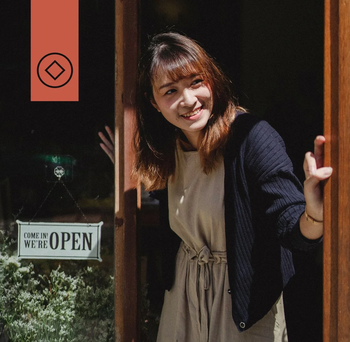 A young woman with brown hair and bangs smiling and peeking out from a wooden door frame at night. There is an