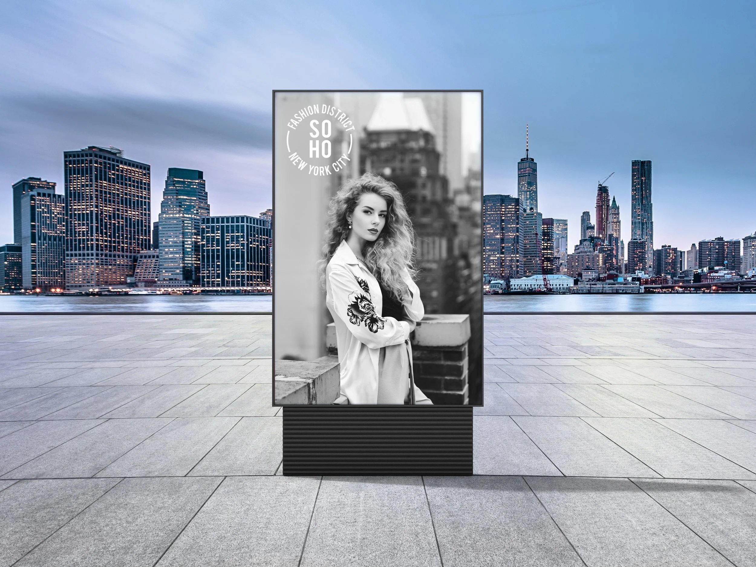 Black and white digital billboard of a woman with curly hair, wearing a jacket with floral embroidery, standing outdoors with city skyscrapers in the background. The poster has a circular logo that reads "Fashion District Soho New York City."