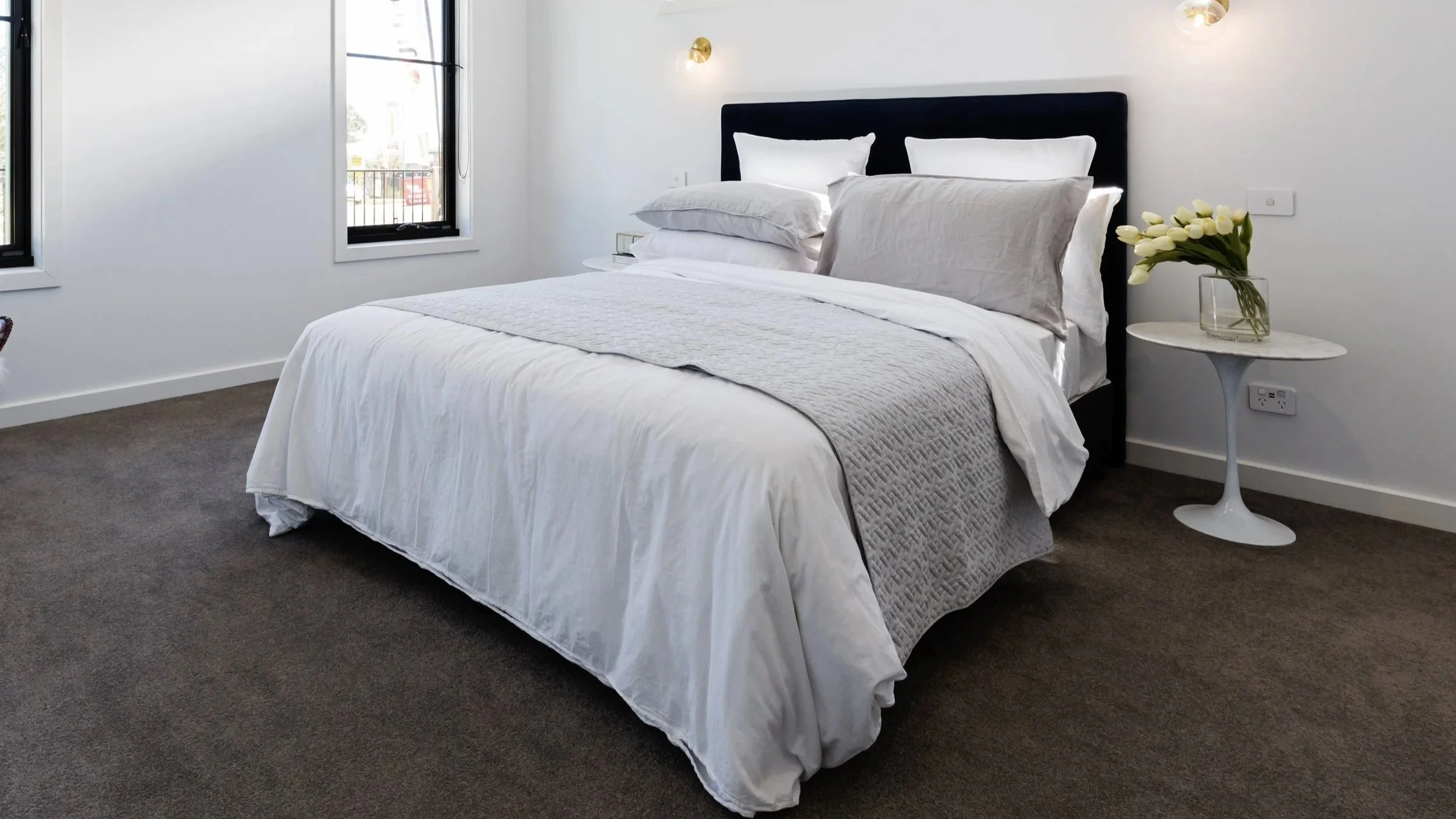 A bedroom with white walls and a dark carpeted floor. There is a bed with white linens and pillows, a black headboard, and a small white side table with a clear glass vase of white flowers. Two wall-mounted lights are above the bed, and three windows