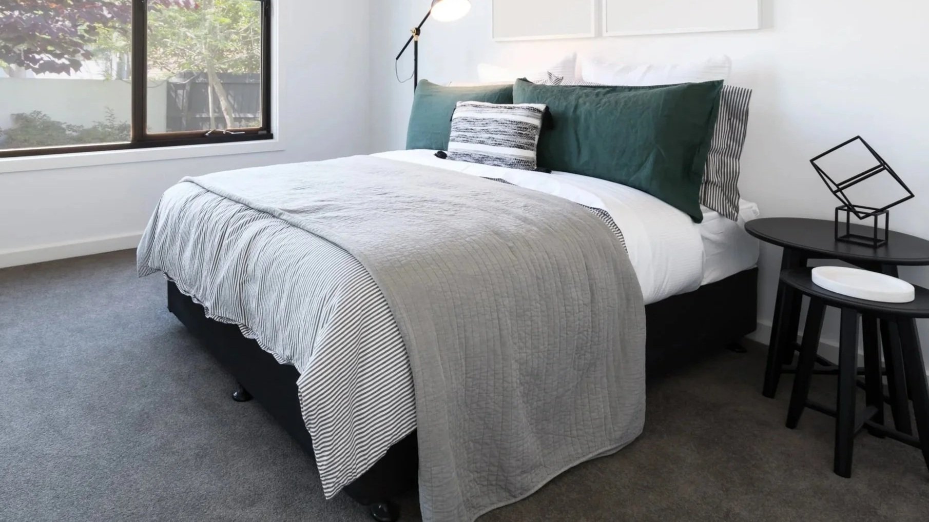 A bedroom with a large bed dressed in black and white bedding, two dark green pillows, and a striped decorative pillow. There is a black side table with geometric decor and a black round table with a white dish. A floor lamp with a gold arm is next t