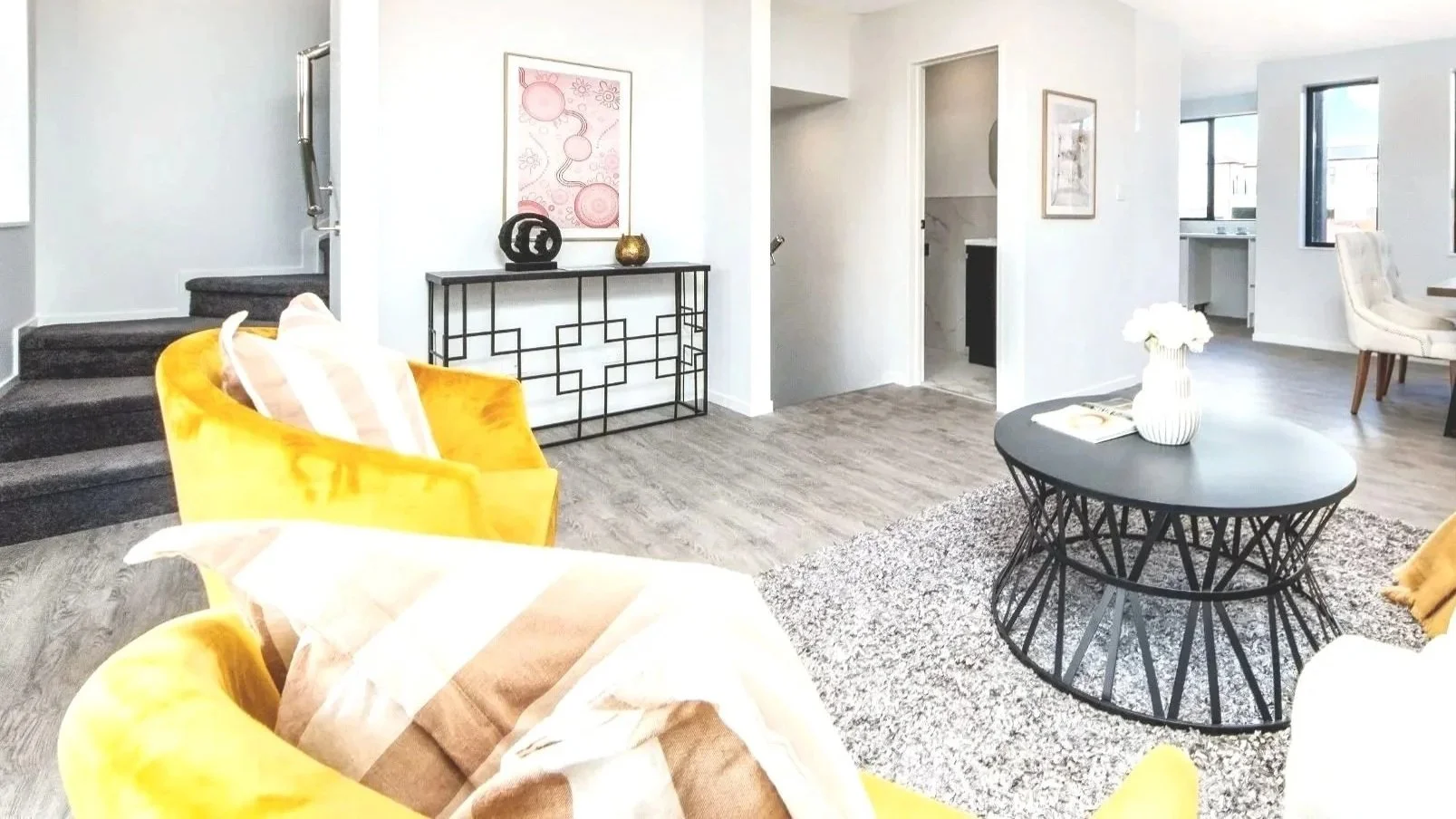 Bright, modern living room with white walls and large windows. Features include a yellow armchair, a black wire coffee table, white dining chairs, and wall art.