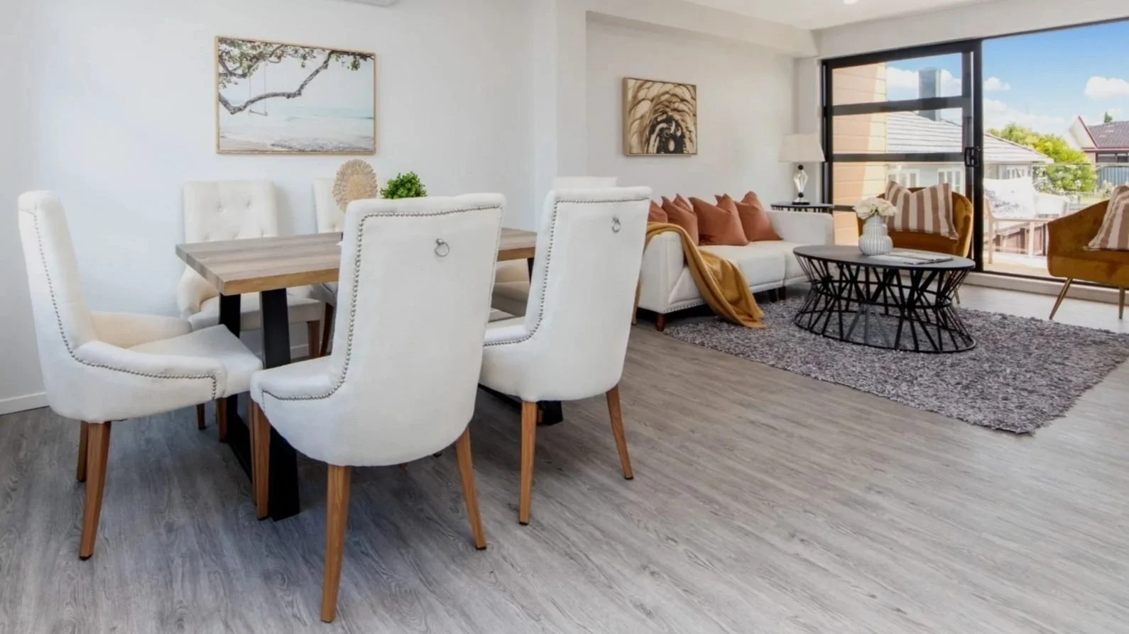 Living and dining room with white walls, large windows, and modern furniture including a dining table with white chairs, a sofa with brown pillows, and wall art.