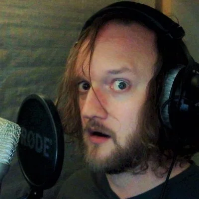 A male voice actor with long hair, beard, and headphones in a recording studio, talking into a microphone.