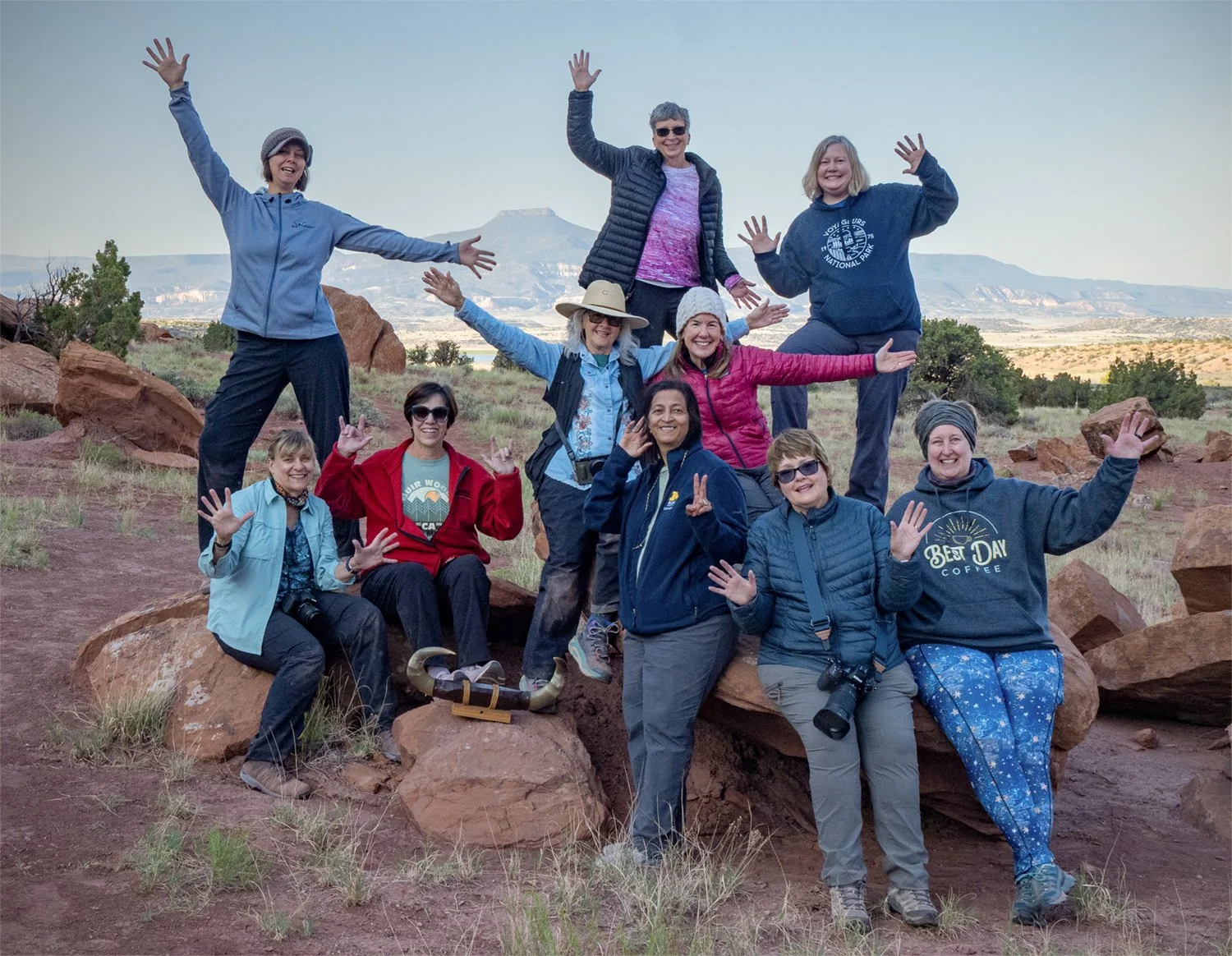 Group of eleven women posing outdoors in a desert landscape with rocks and distant mountains, some standing on rocks, making gestures with their hands, smiling, and wearing casual outdoor clothing and hats.