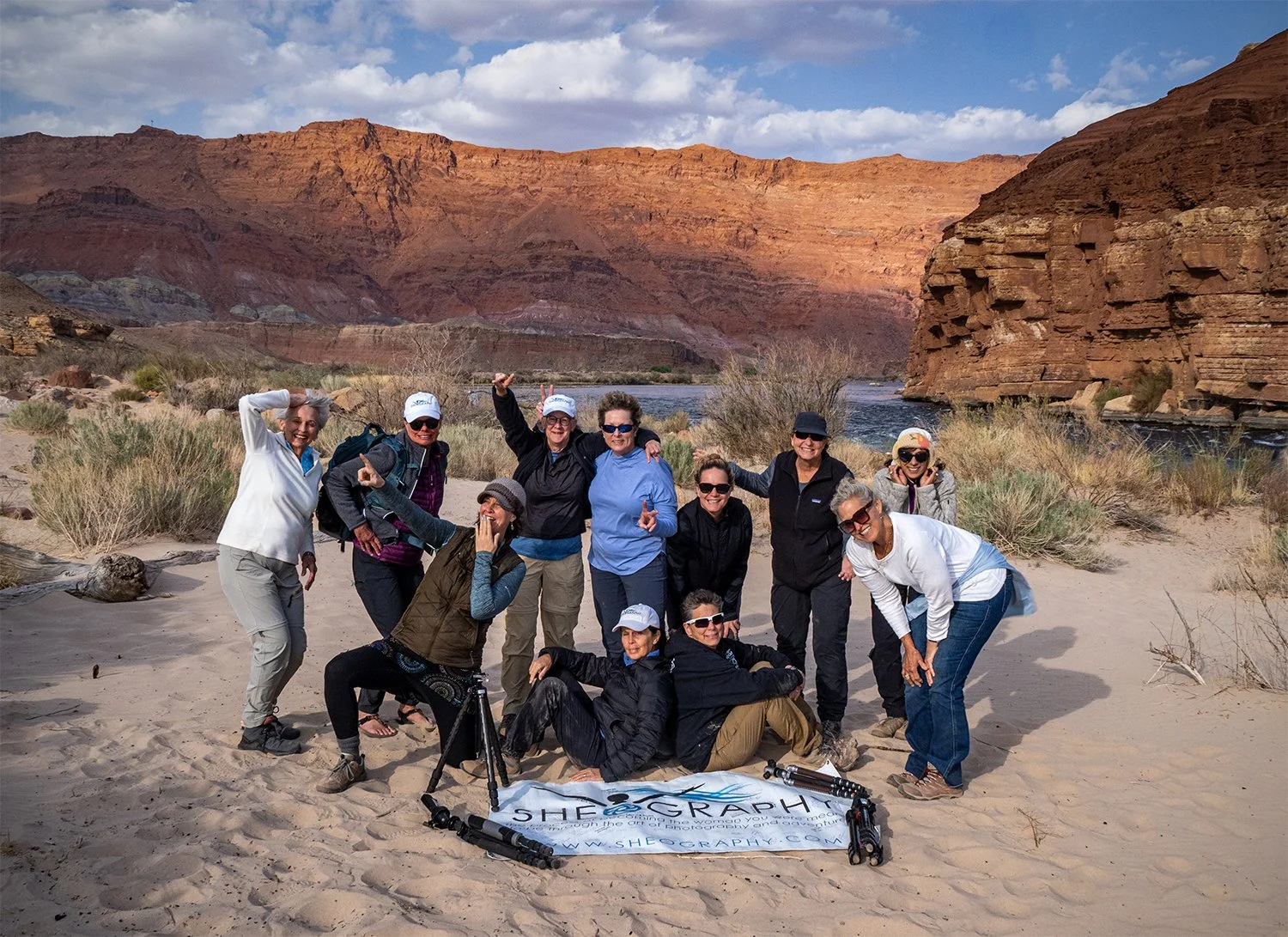Group of eleven people smiling and posing on a sandy riverbank with red rock cliffs in the background, holding a banner that reads 'SHEOGRAPHY'.