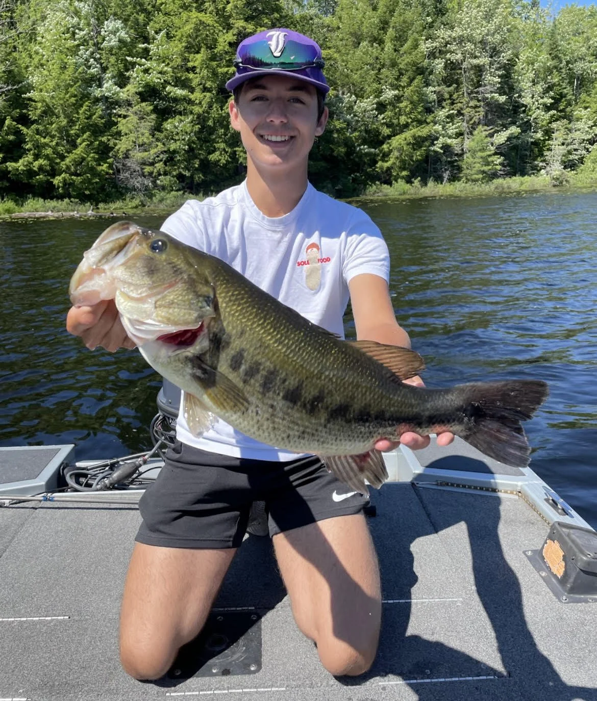 Person holding a large fish on a boat in a lake, surrounded by trees.
