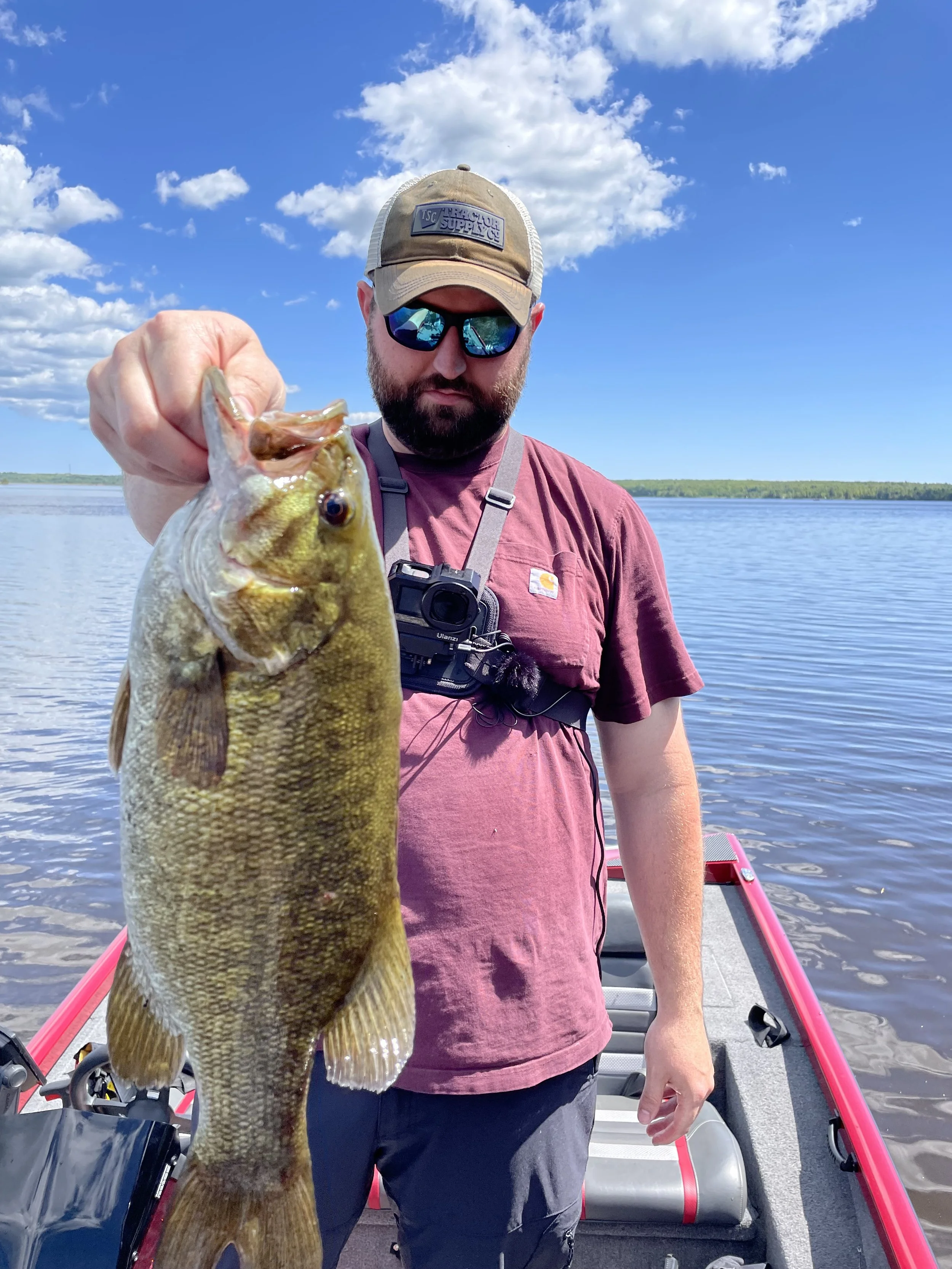 Person holding a largemouth bass fish on a boat, wearing a cap and sunglasses, with a lake and blue sky in the background.