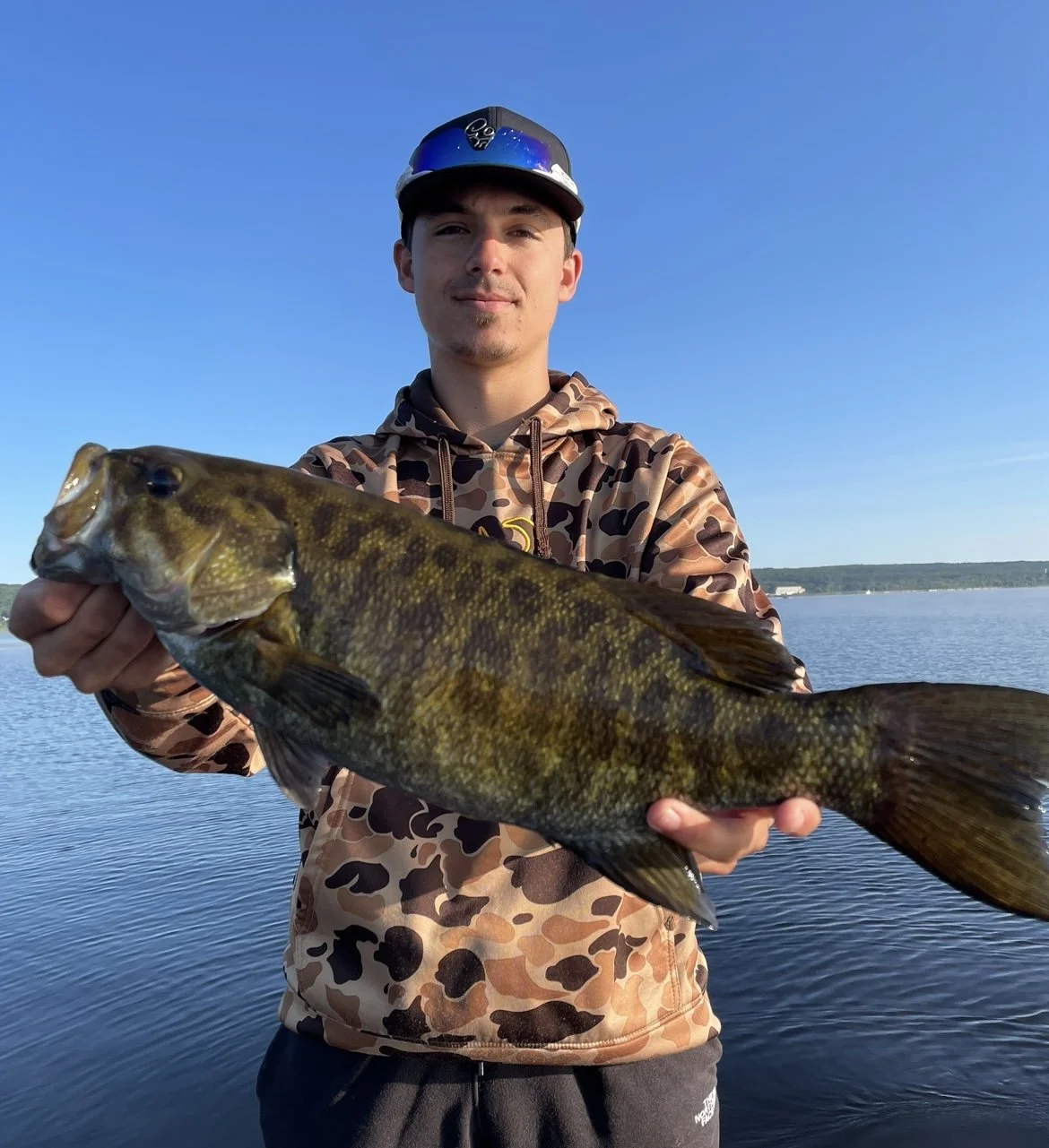 Person holding a largemouth bass near a lake in a camo hoodie and cap.