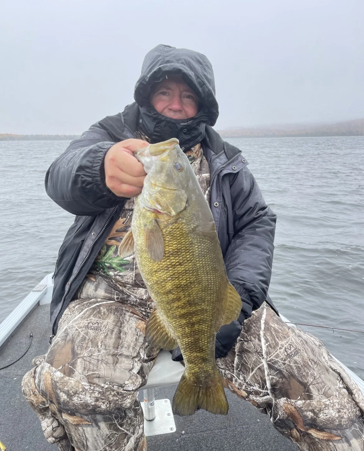 Person in camo clothing holding a large fish on a boat in a lake.