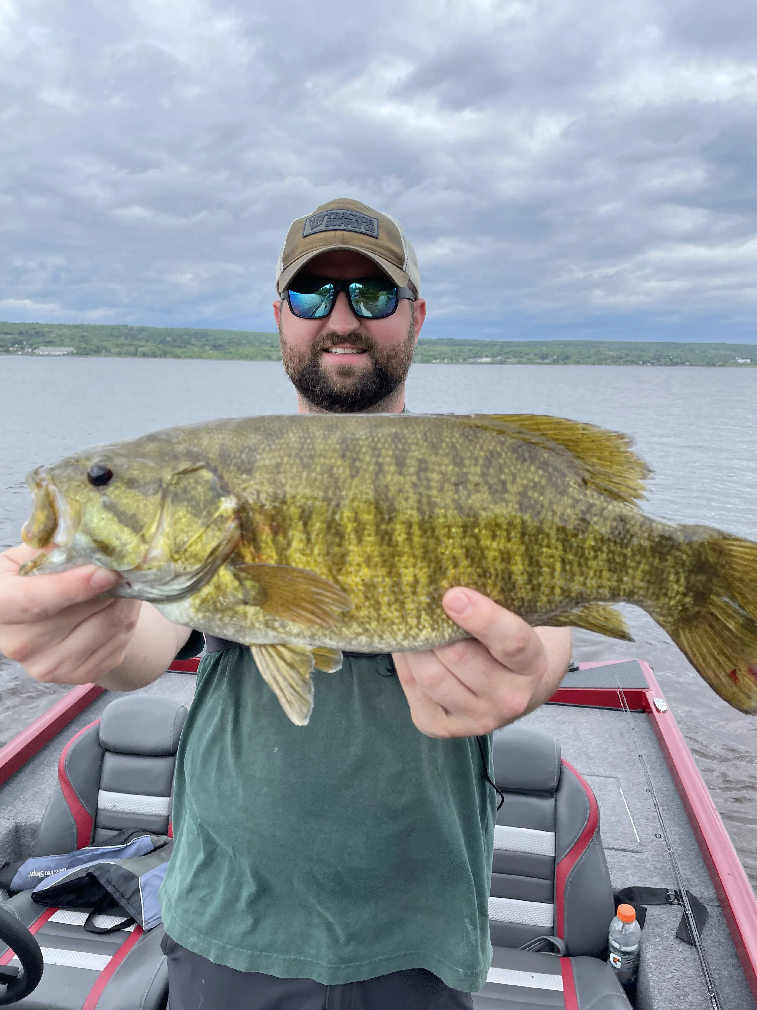 Person holding a large fish on a boat with a lake and cloudy sky in the background.