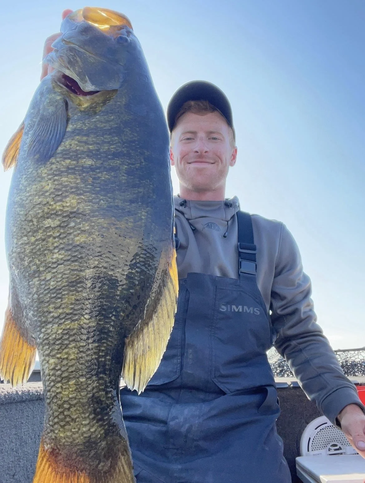 Man holding a large fish on a boat, wearing a grey jacket and cap, smiling.