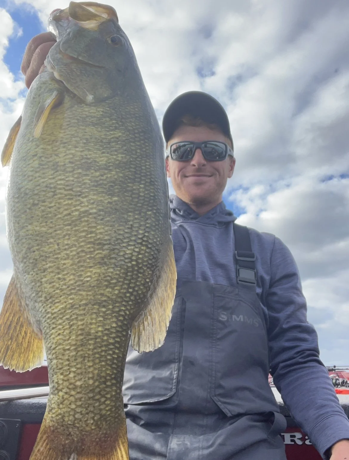 Person holding a large fish, wearing sunglasses and a cap on a cloudy day.