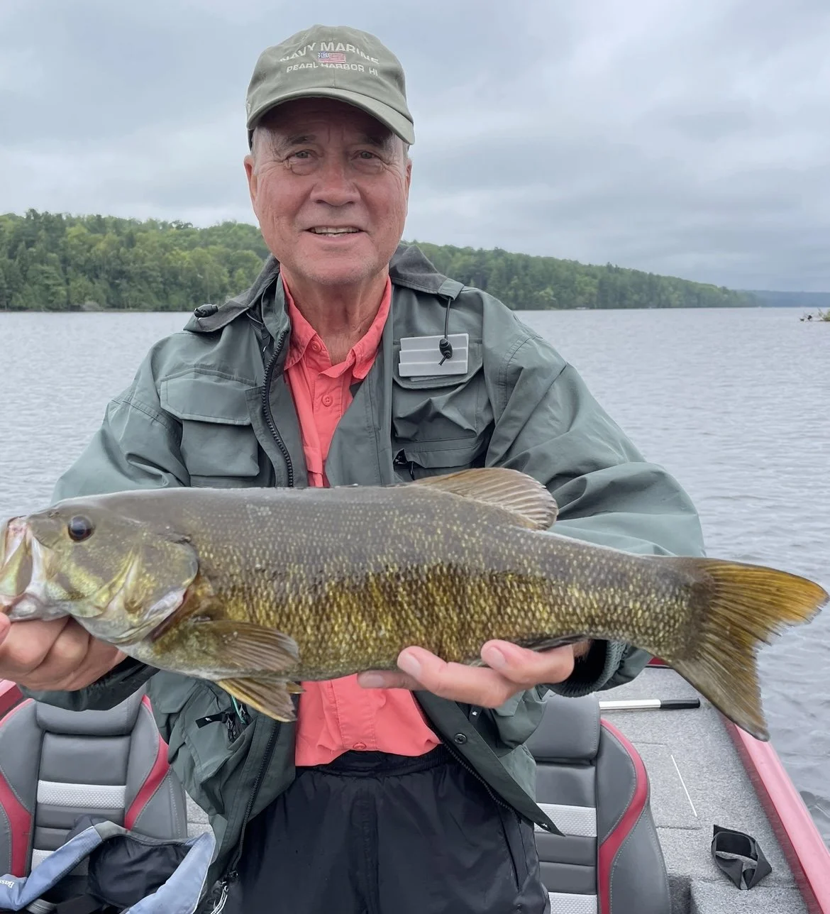 Bass Fishing Guide Services - Fishing Guide - Chassell, MI - Man holding a bass fish on a boat with a lake in the background.