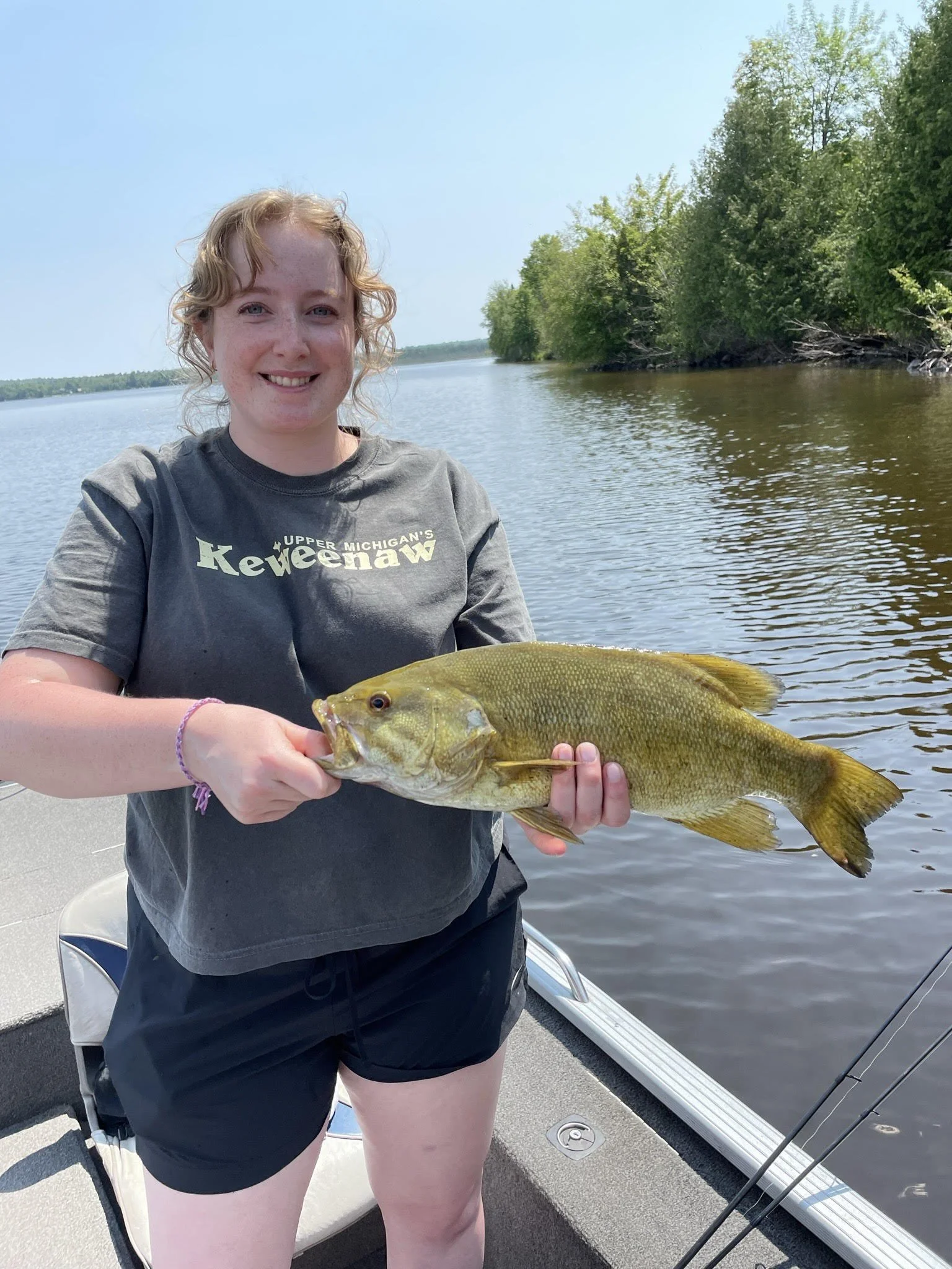 Person holding a fish on a boat near a lake shore with trees in the background.