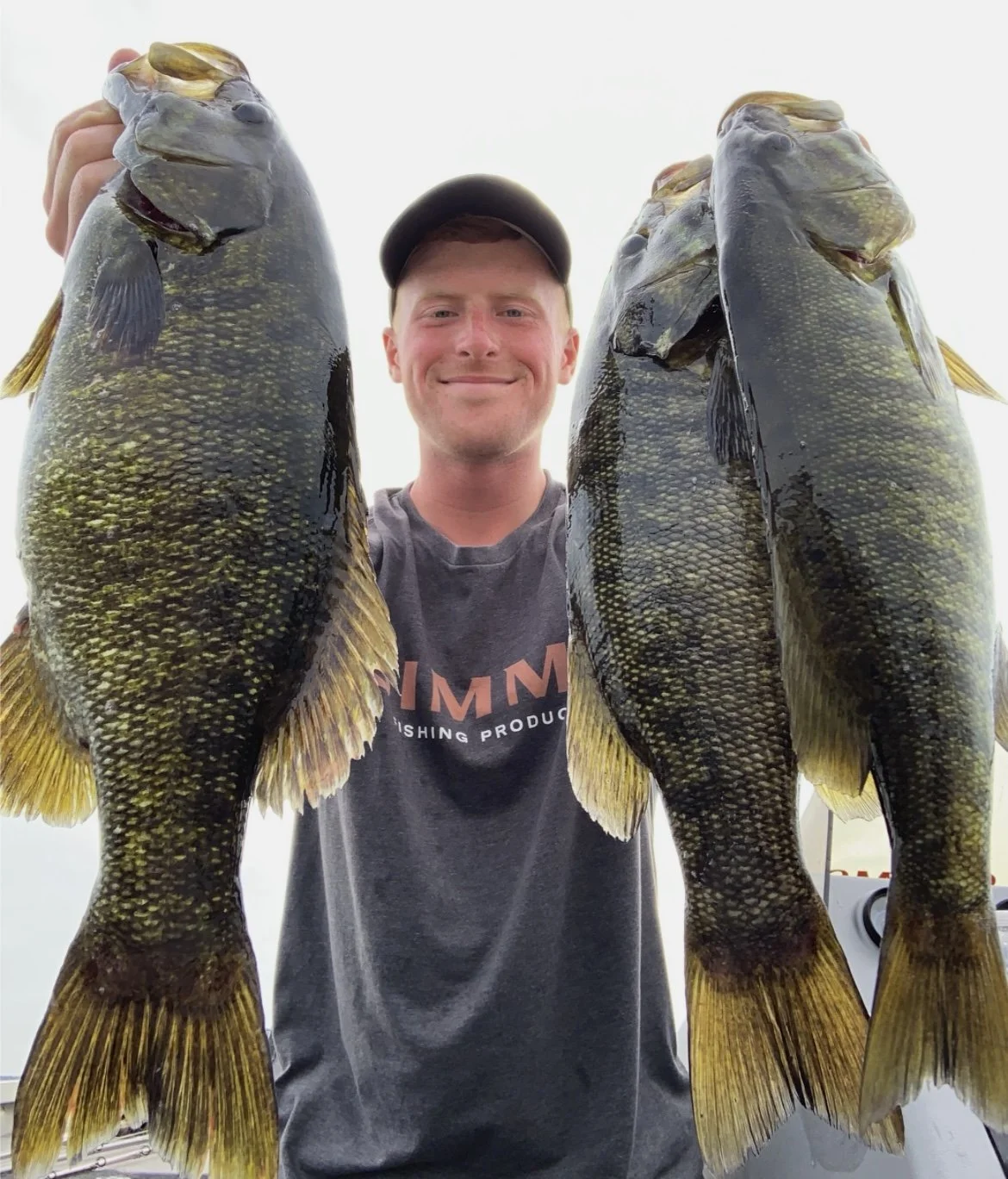 Man holding three large fish, smiling, wearing a cap and a dark T-shirt with "SIMM" visible. The fish appear to be bass.