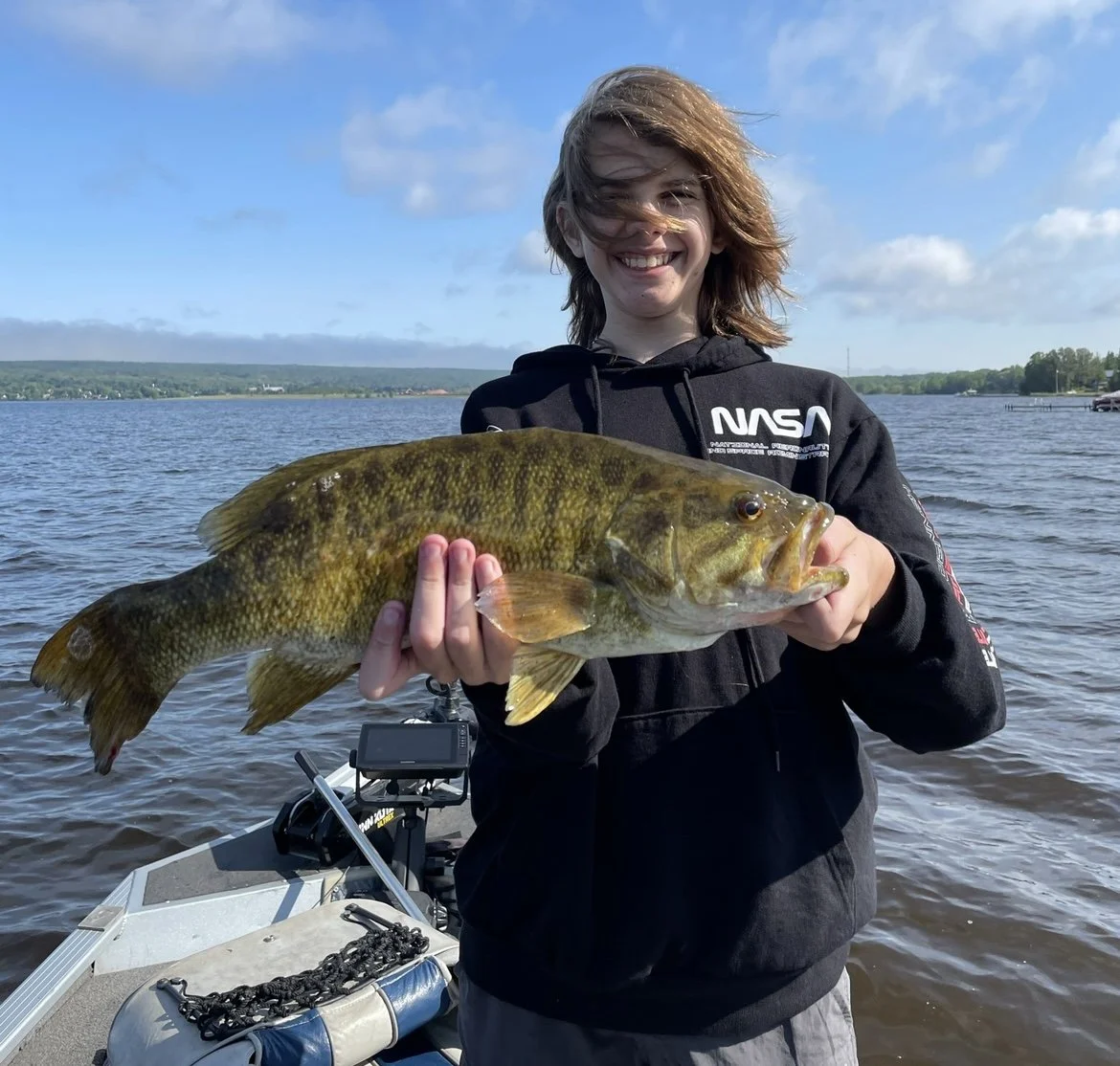 Person holding a large fish on a boat by a lake.