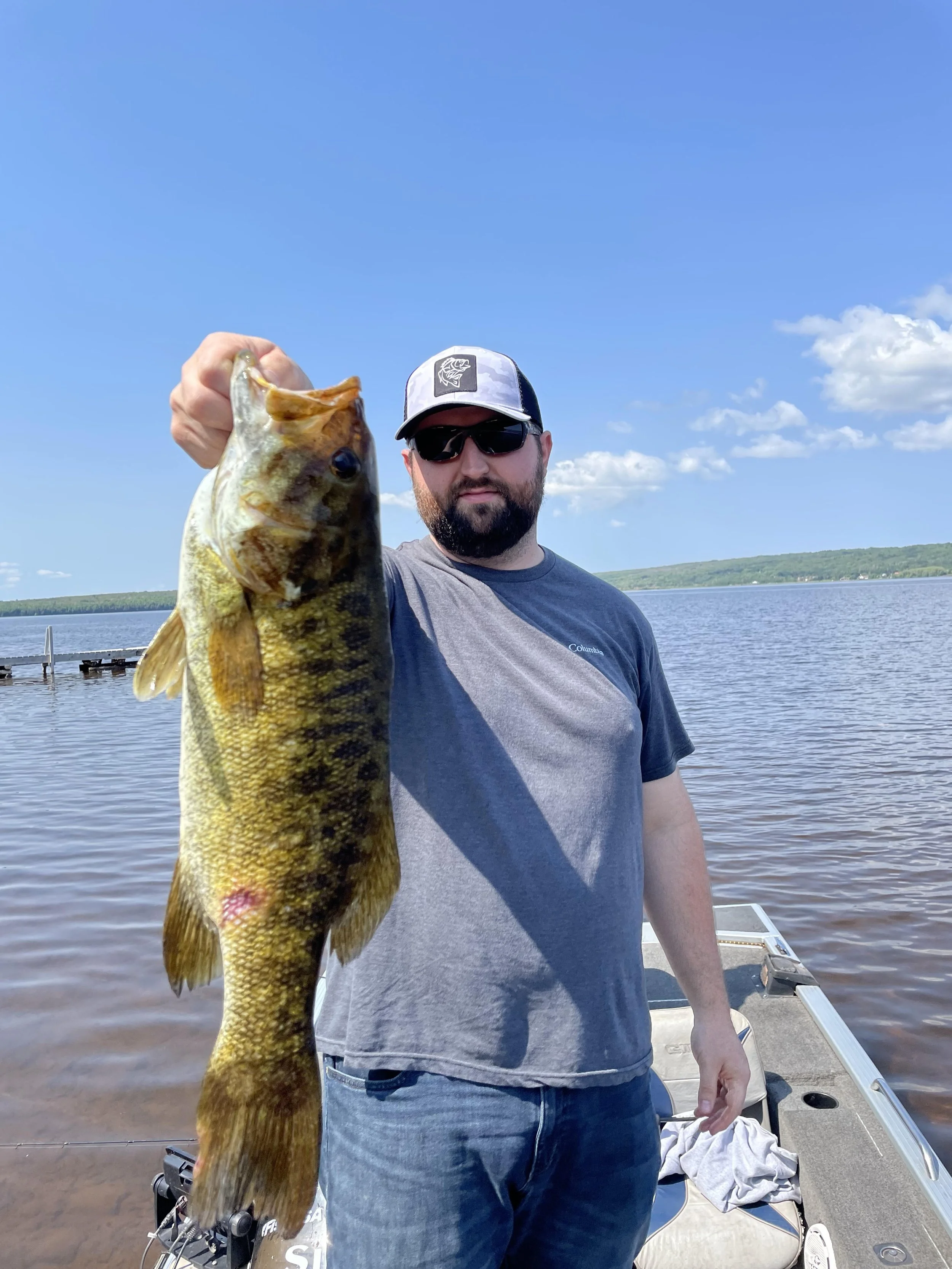 Man holding a large fish on a boat in a lake under a clear blue sky.