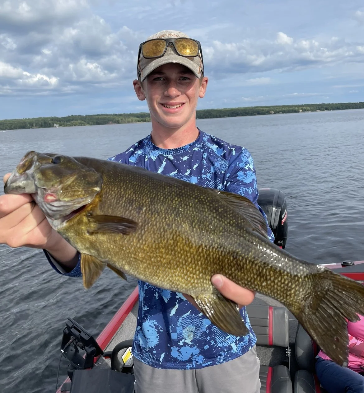 Person holding a large fish in front of a lake, wearing a blue camouflage shirt and a cap.